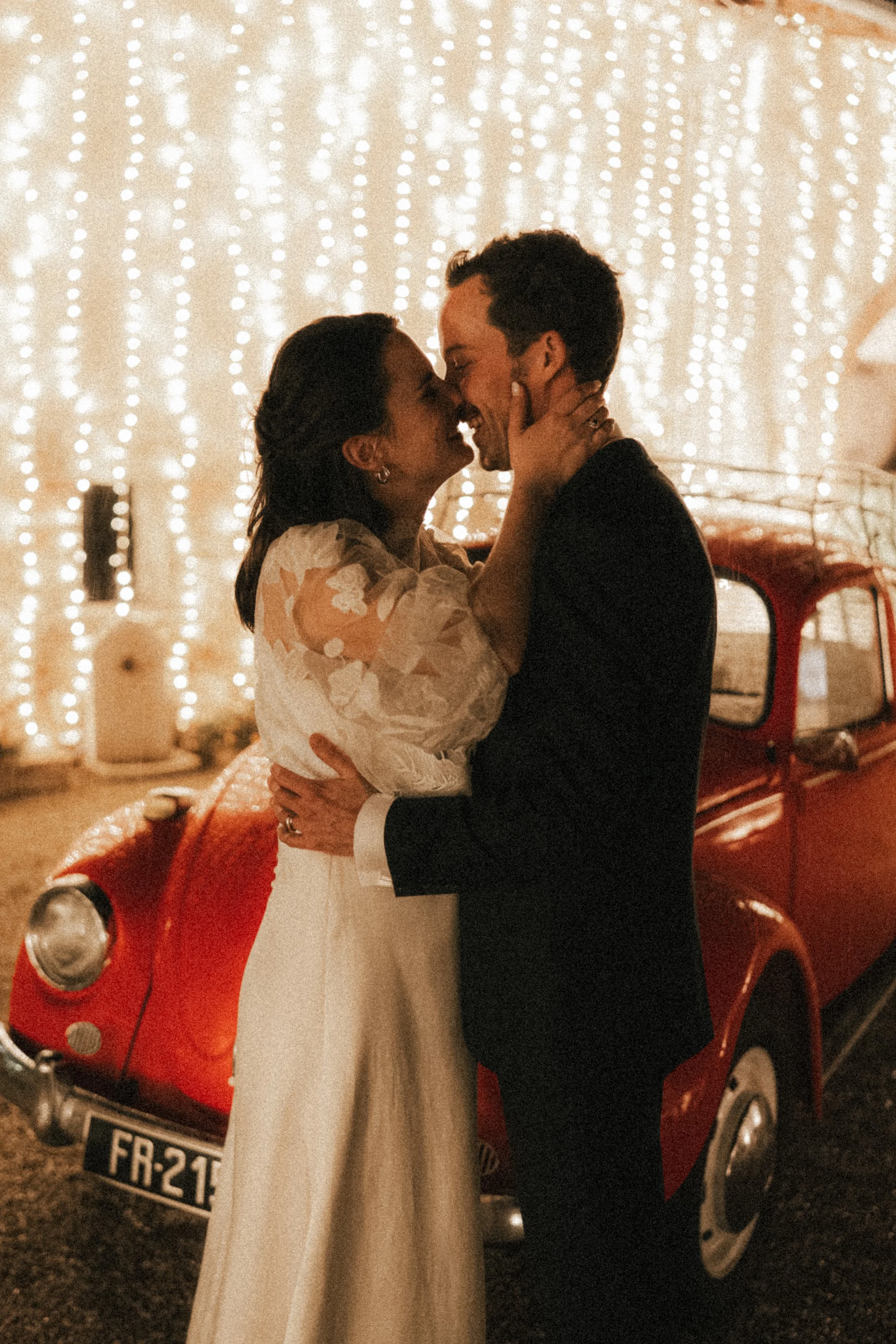 Un couple s'embrasse lors d'une occasion spéciale, avec des guirlandes lumineuses en arrière-plan et une voiture rouge vintage.
