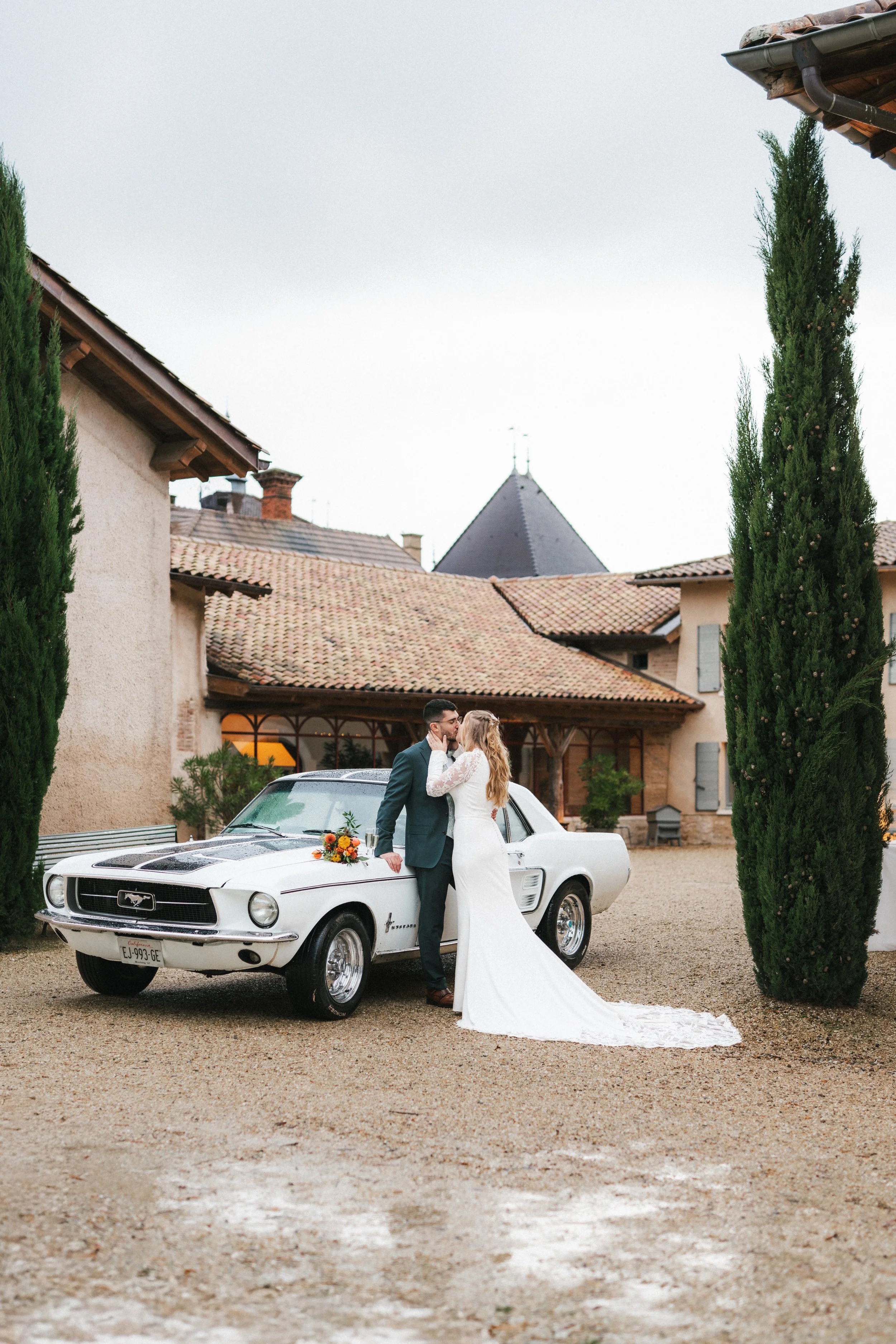 Un couple de mariés s'embrassant devant une voiture de collection blanche dans une cour de village avec des maisons en pierre et deux grands cèdres.