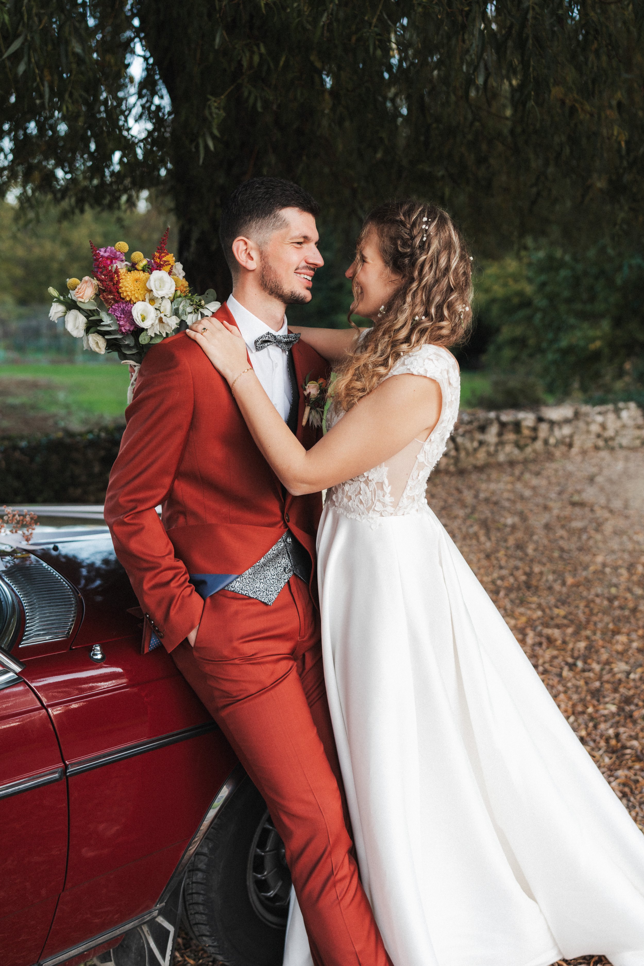 Un couple en robe de mariée et costume, souriant, se tenant proche devant une voiture rouge en pleine nature, lors d'un mariage en plein air.