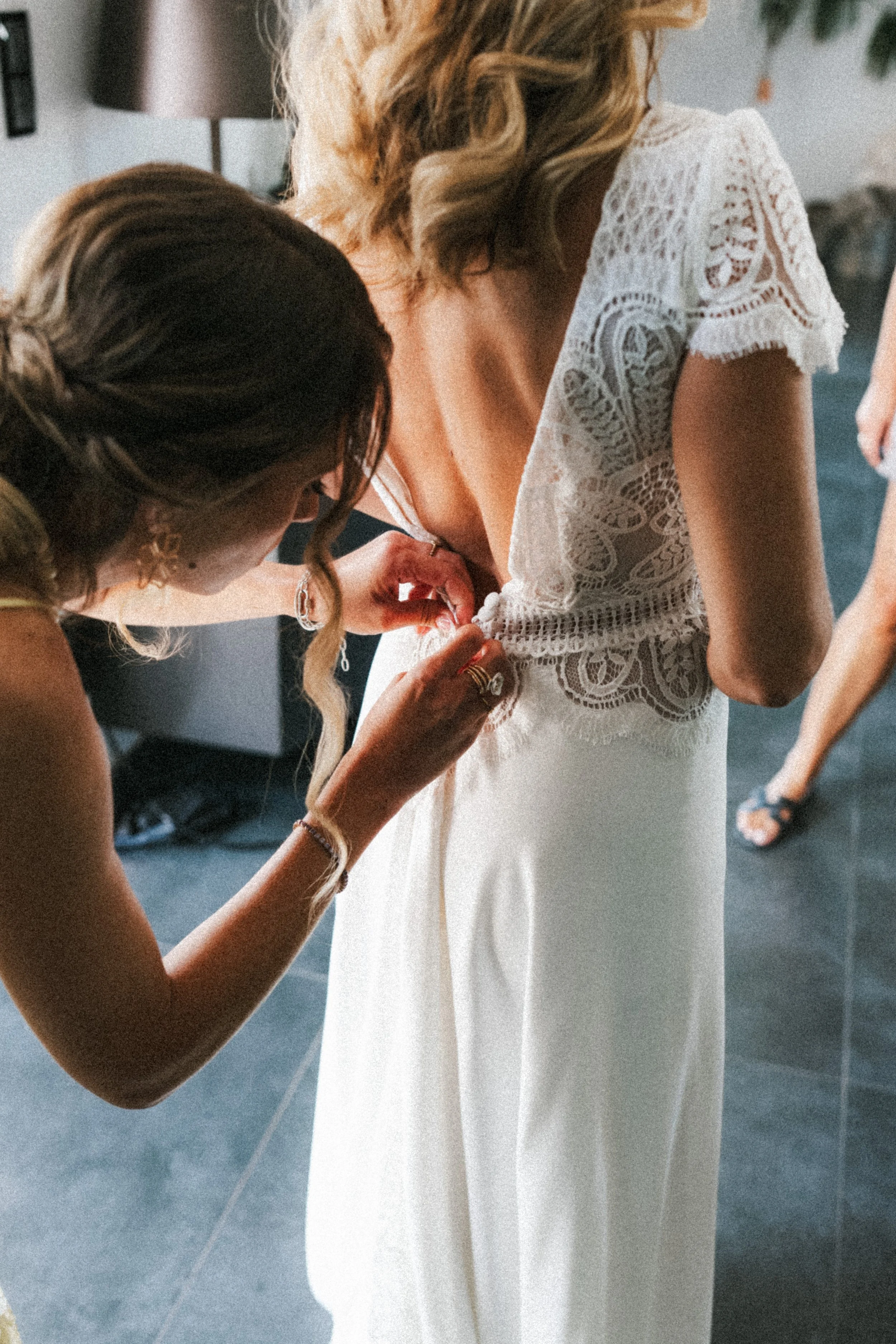 Une femme en robe blanche avec des détails en dentelle est aidée à boutonner sa robe par une autre femme. La femme en robe a des cheveux blonds, ondulés.