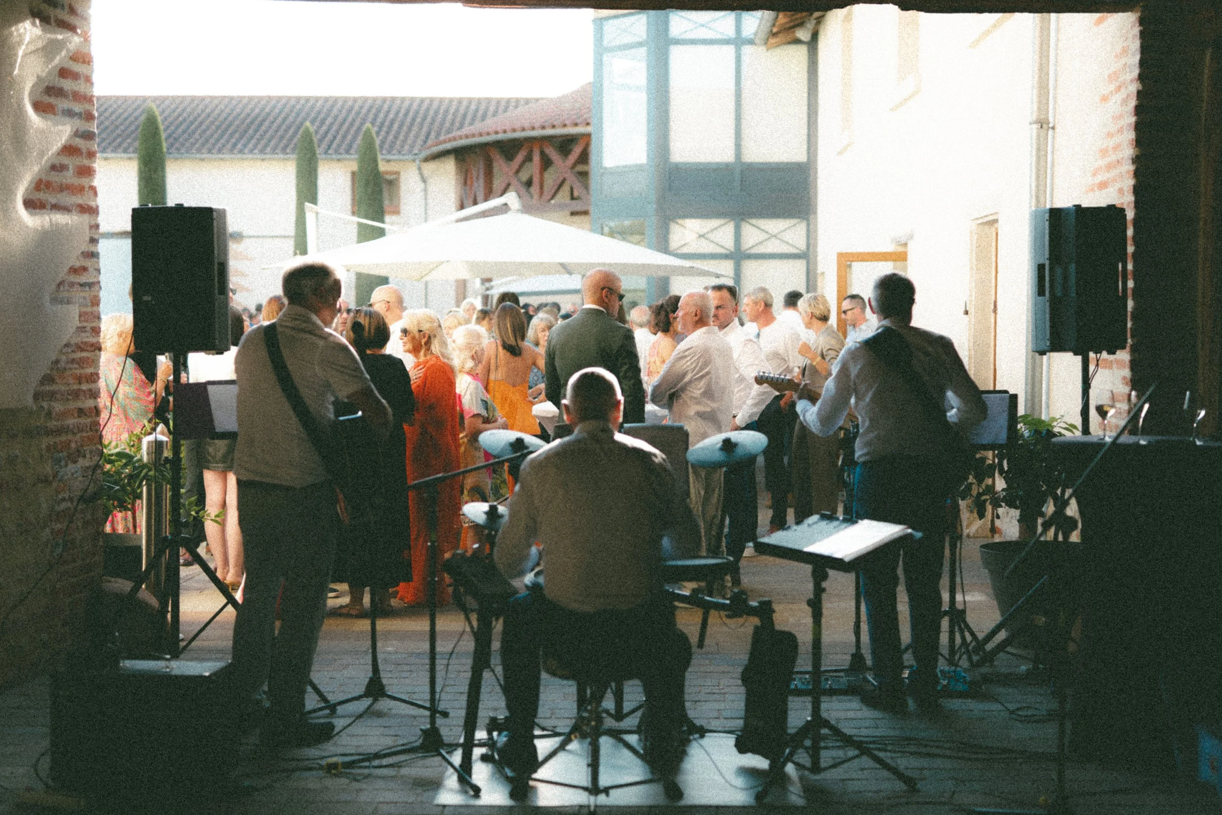Un groupe de musiciens joue lors d'une fête en intérieur/exérieur avec un grand groupe de personnes qui dansent et socialisent sous un parasol