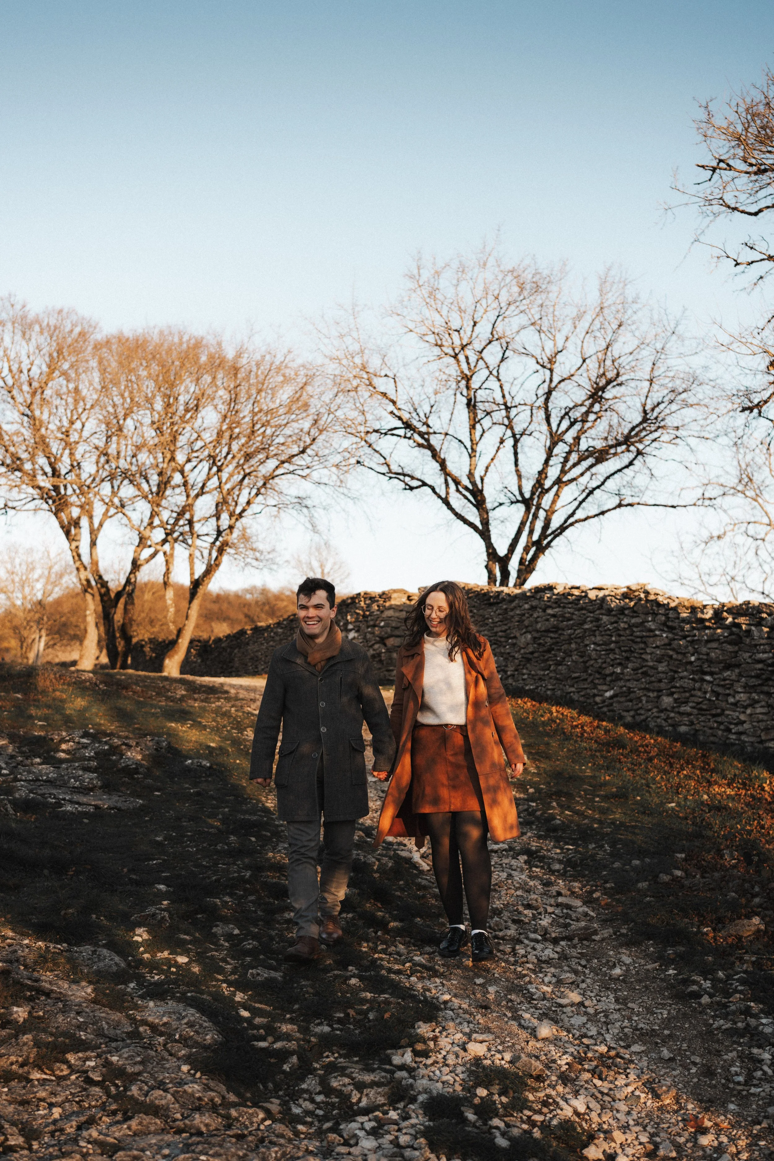 Un couple marche main dans la main dans un paysage en automne, avec des arbres sans feuilles et un mur en pierres en arrière-plan, sous un ciel bleu clair.