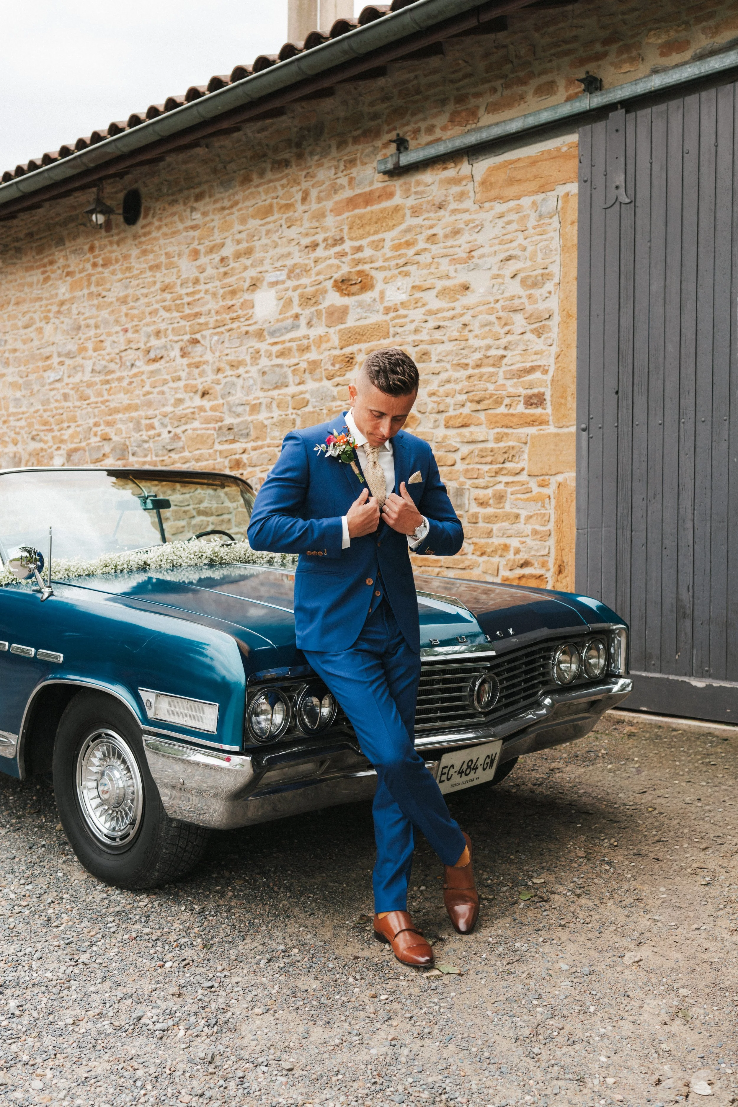 Jeune homme en costume bleu, regardant vers le sol, devant une voiture ancienne décorée avec des fleurs, probablement à un mariage.