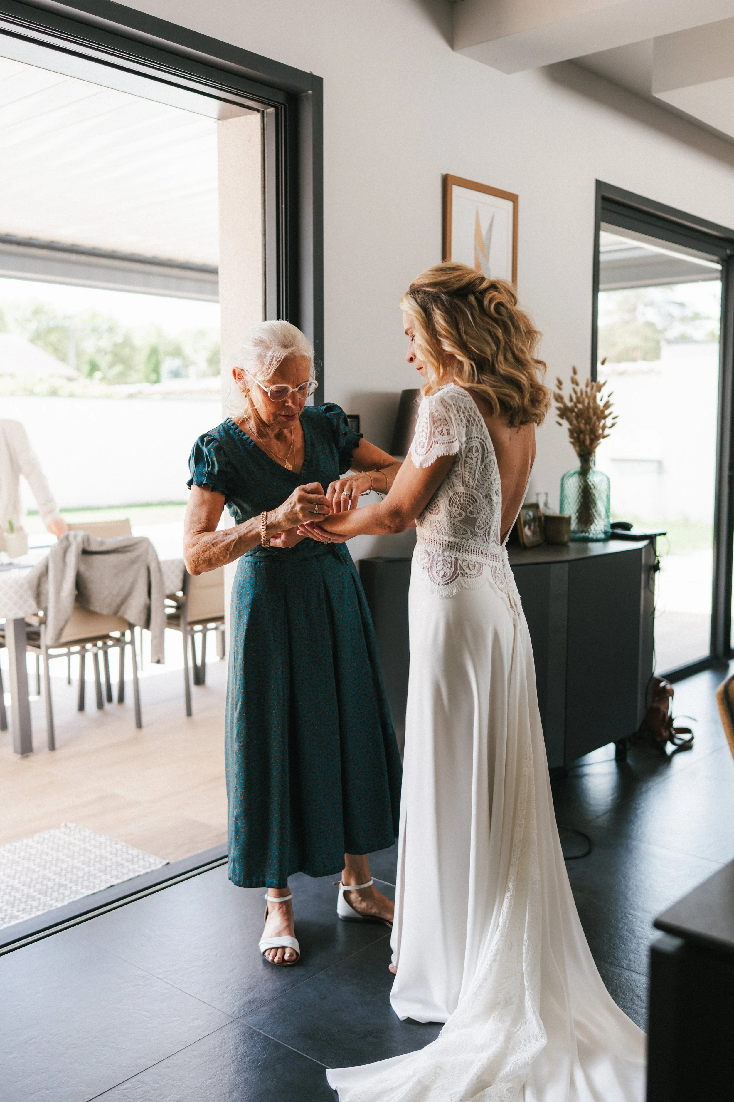 Une jeune femme en robe de mariée aide une femme âgée à mettre une bague. La scène se déroule dans une maison lumineuse avec des fenêtres grandes ouvertes, décorée simplement avec un meuble noir et une plante en vase.