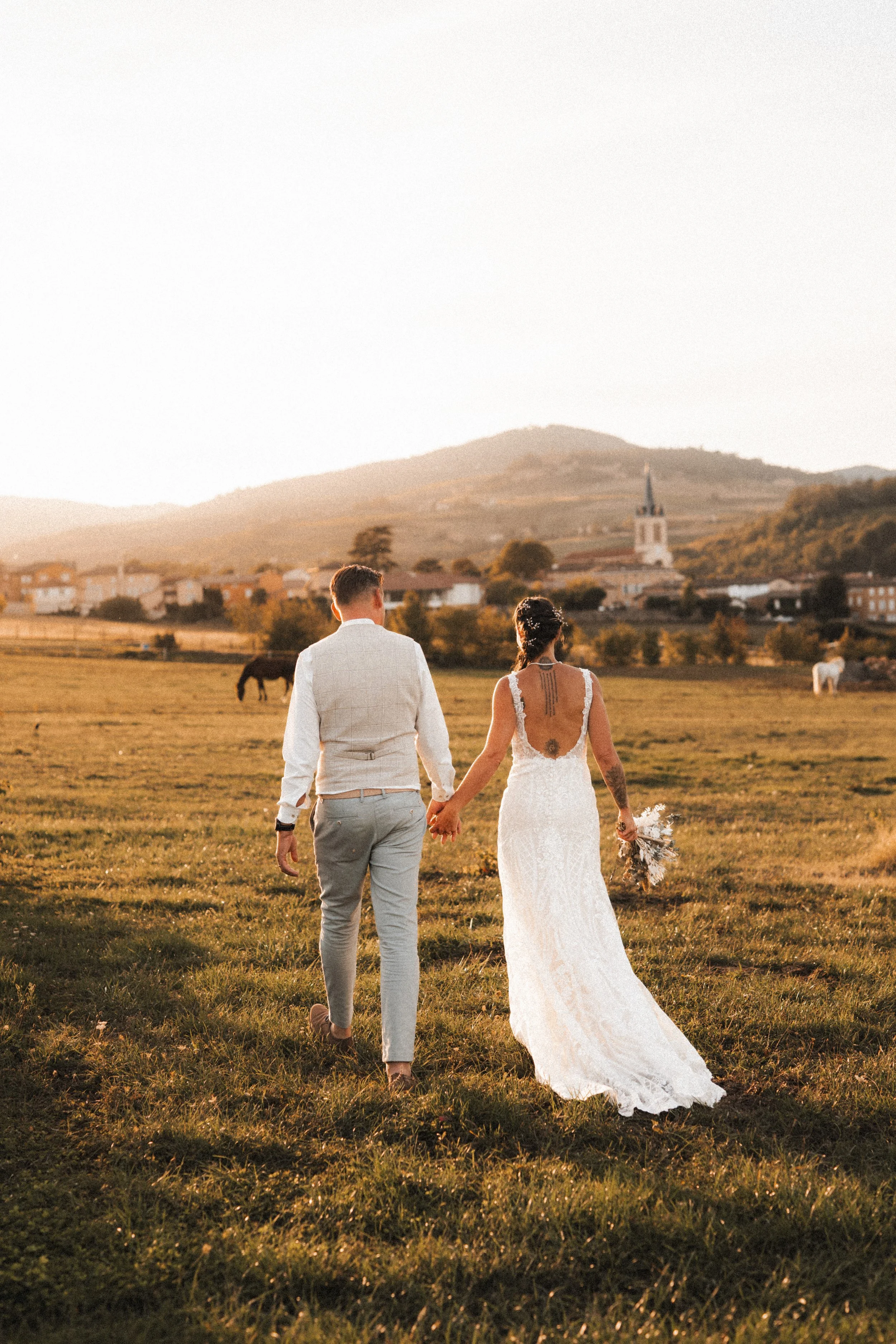 Un couple en mariage marche dans un champ lors d'un coucher de soleil, tenant la main avec une église en arrière-plan dans un paysage rural.