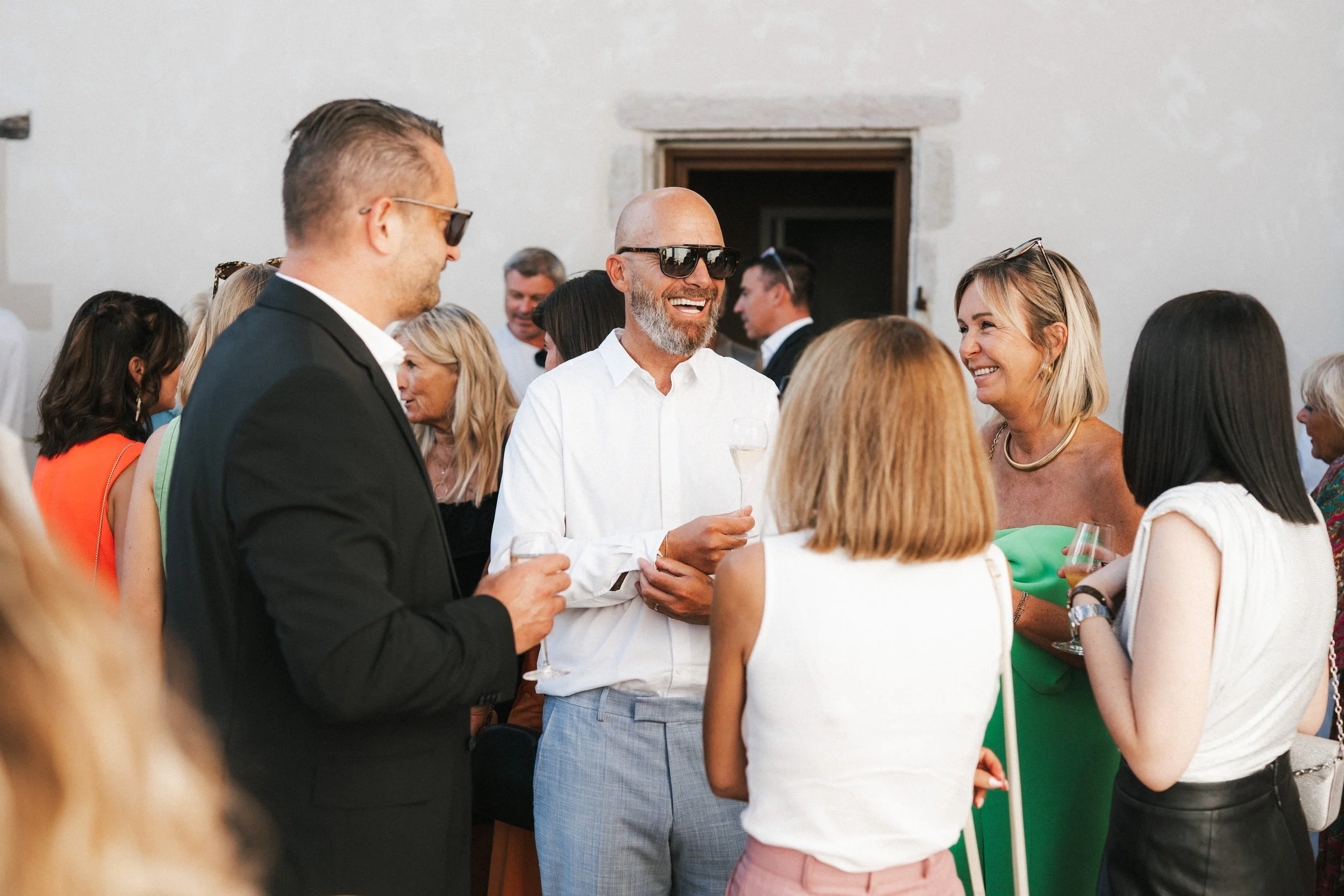 Groupe de personnes lors d'une réception ou d'une fête, portant des vêtements élégants, certaines avec des lunettes de soleil, en train de discuter et de sourire, en intérieur ou en extérieur