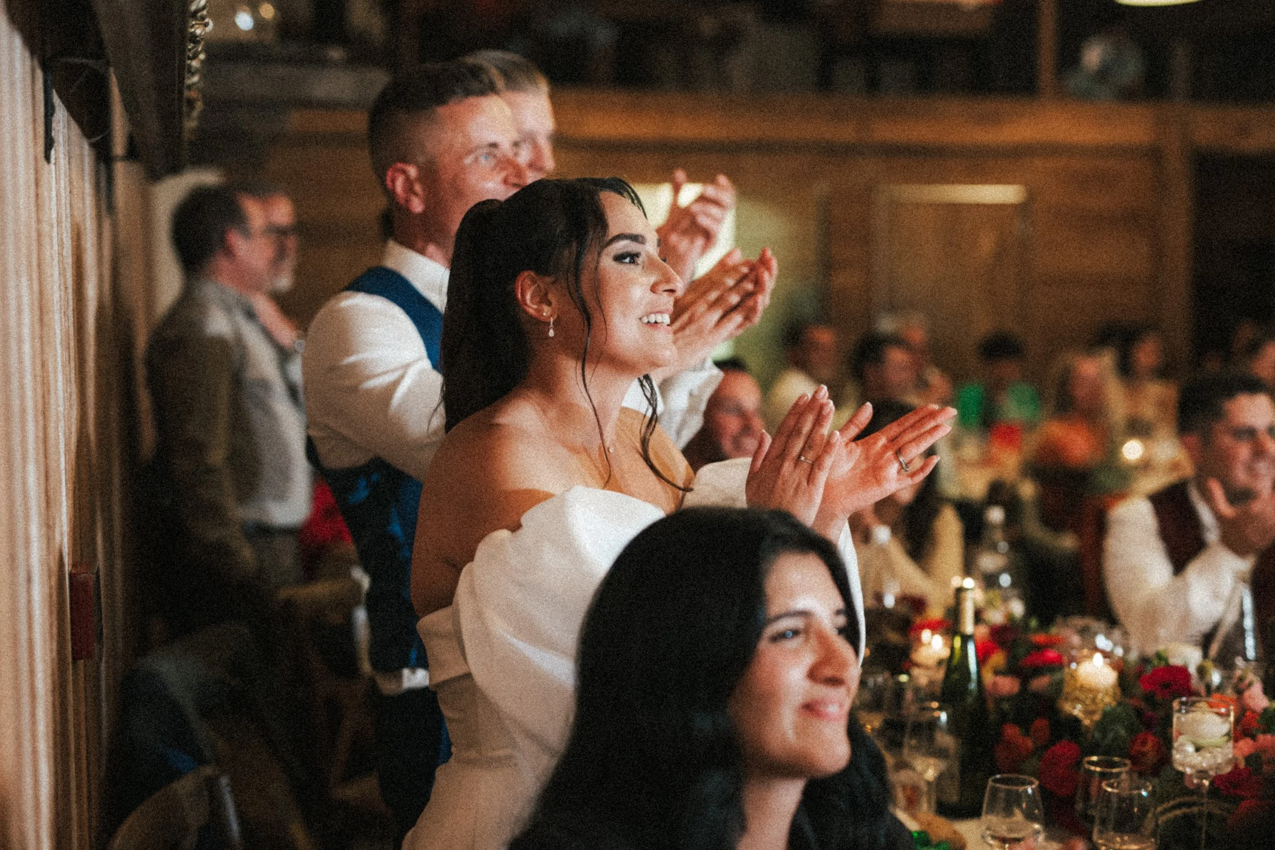 Un groupe de personnes lors d'une célébration, probablement un mariage, assises à une table décorée avec des fleurs et des bougies, applaudissant et souriant.