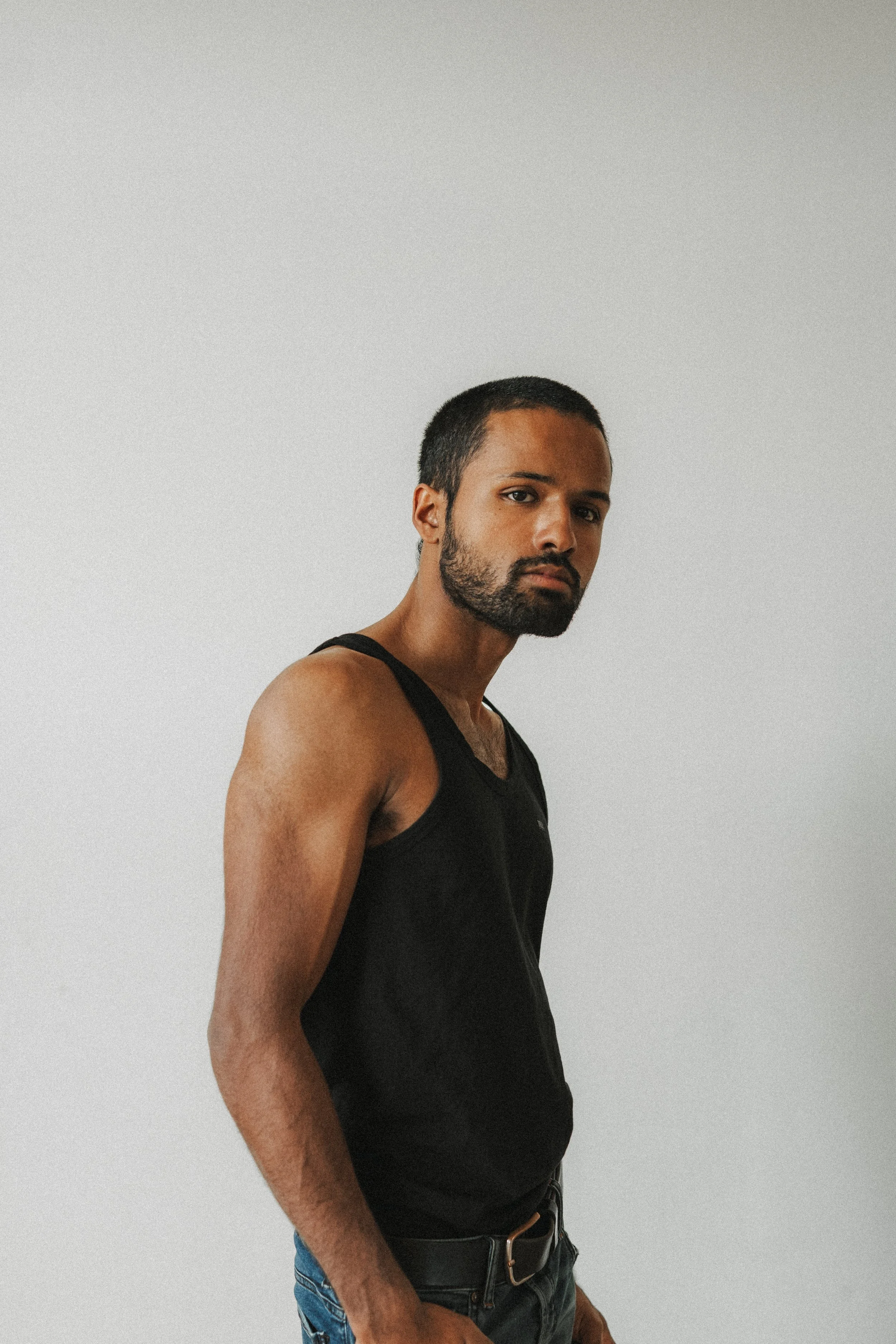Un homme avec une barbe, portant un débardeur noir, pose devant un mur blanc.