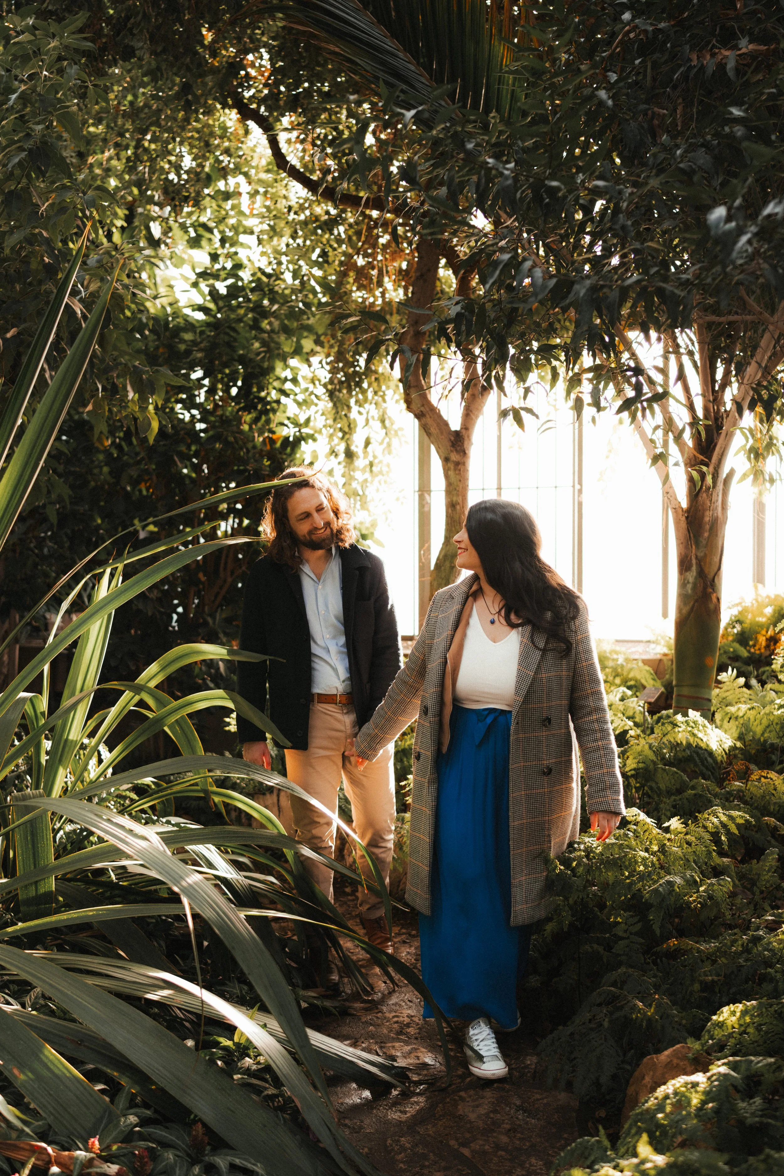Un homme et une femme se tenant la main dans une serre remplie de plantes vertes, avec de la lumière du soleil filtrant à travers les fenêtres.