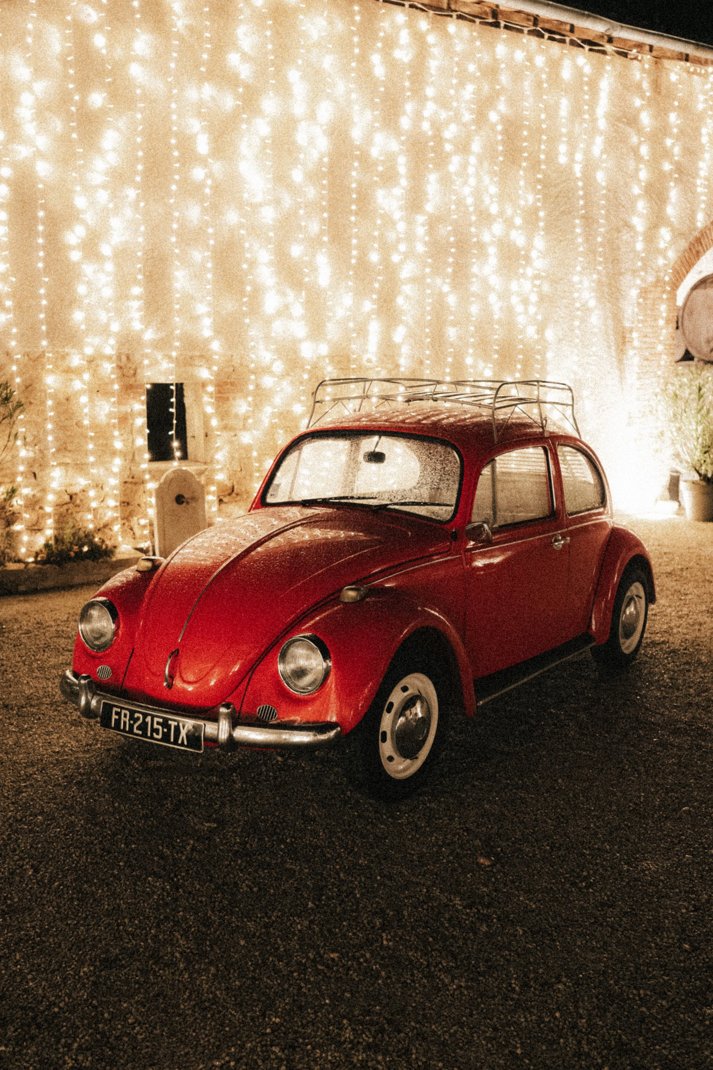 Voiture rouge vintage type Volkswagen Beetle garée devant un mur éclairé de guirlandes lumineuses lors d'une soirée