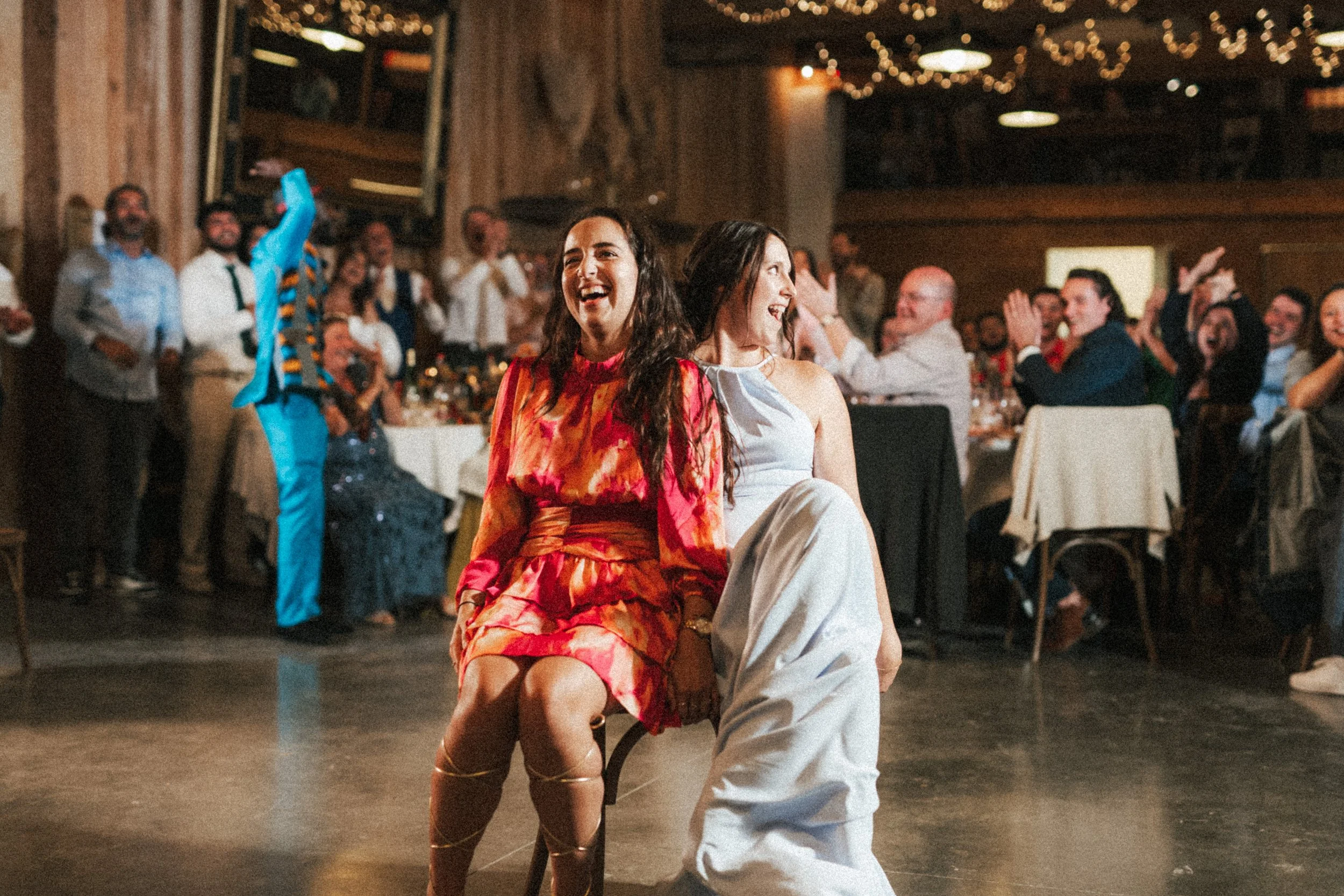 Deux femmes dans une robe de soirée dansant lors d'une célébration, avec un groupe de personnes souriantes assises et debout en arrière-plan dans une salle décorée avec des lumières