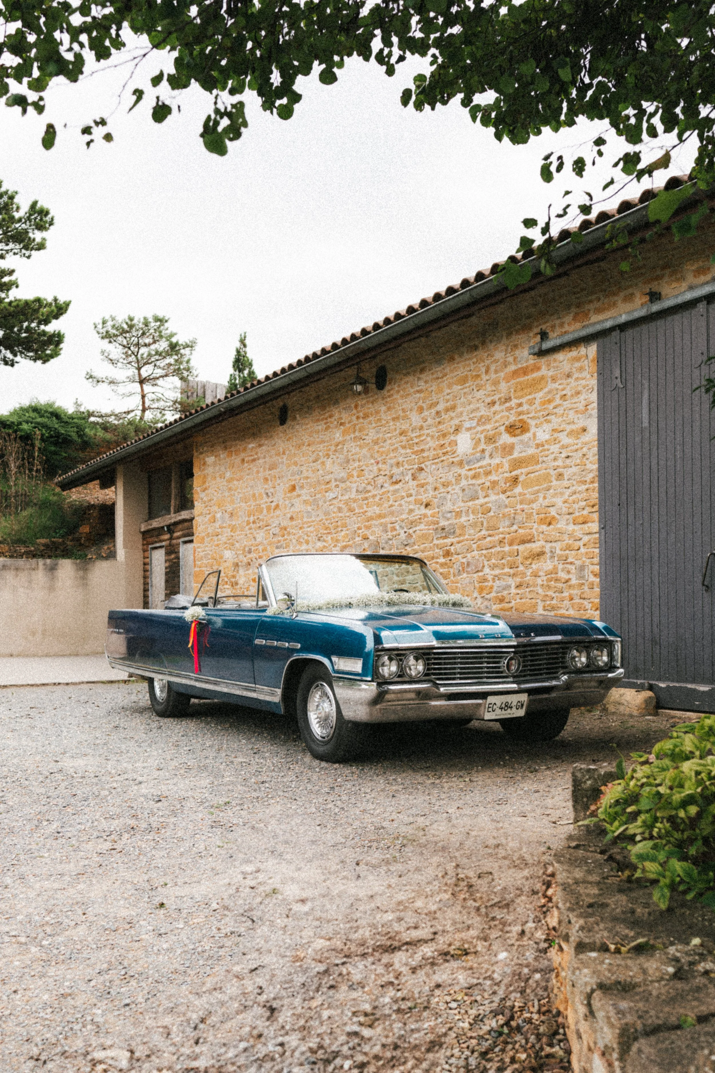 Une vieille voiture cabriolet bleue stationnée devant une maison en pierre avec un toit en tuiles, entourée d'arbres et de végétation, sous un ciel nuageux.