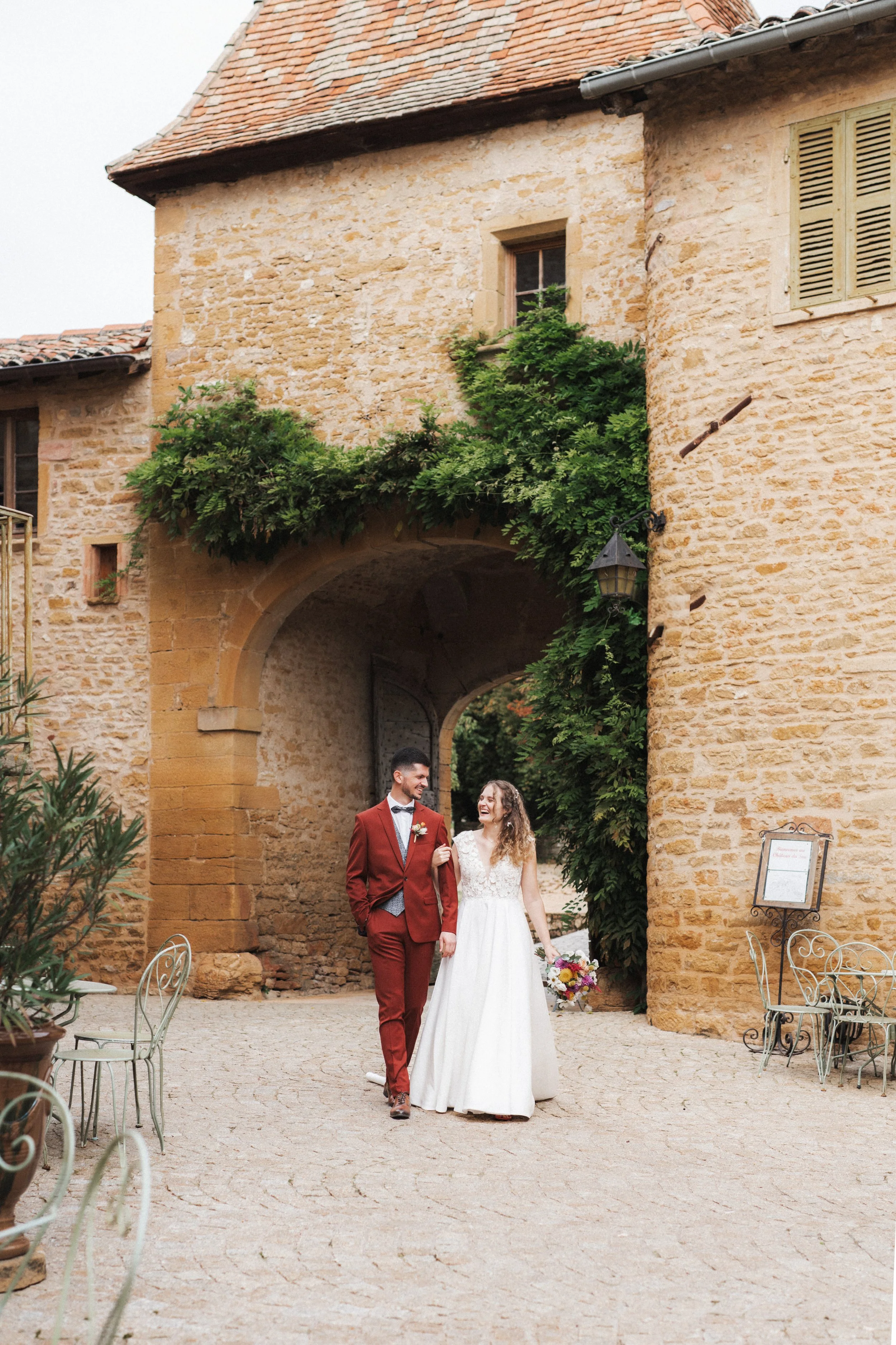 Un couple marié se promenant dans une cour en pierre avec un bâtiment en pierre en arrière-plan, entouré de végétation et de mobilier en fer forgé, lors d'un mariage.