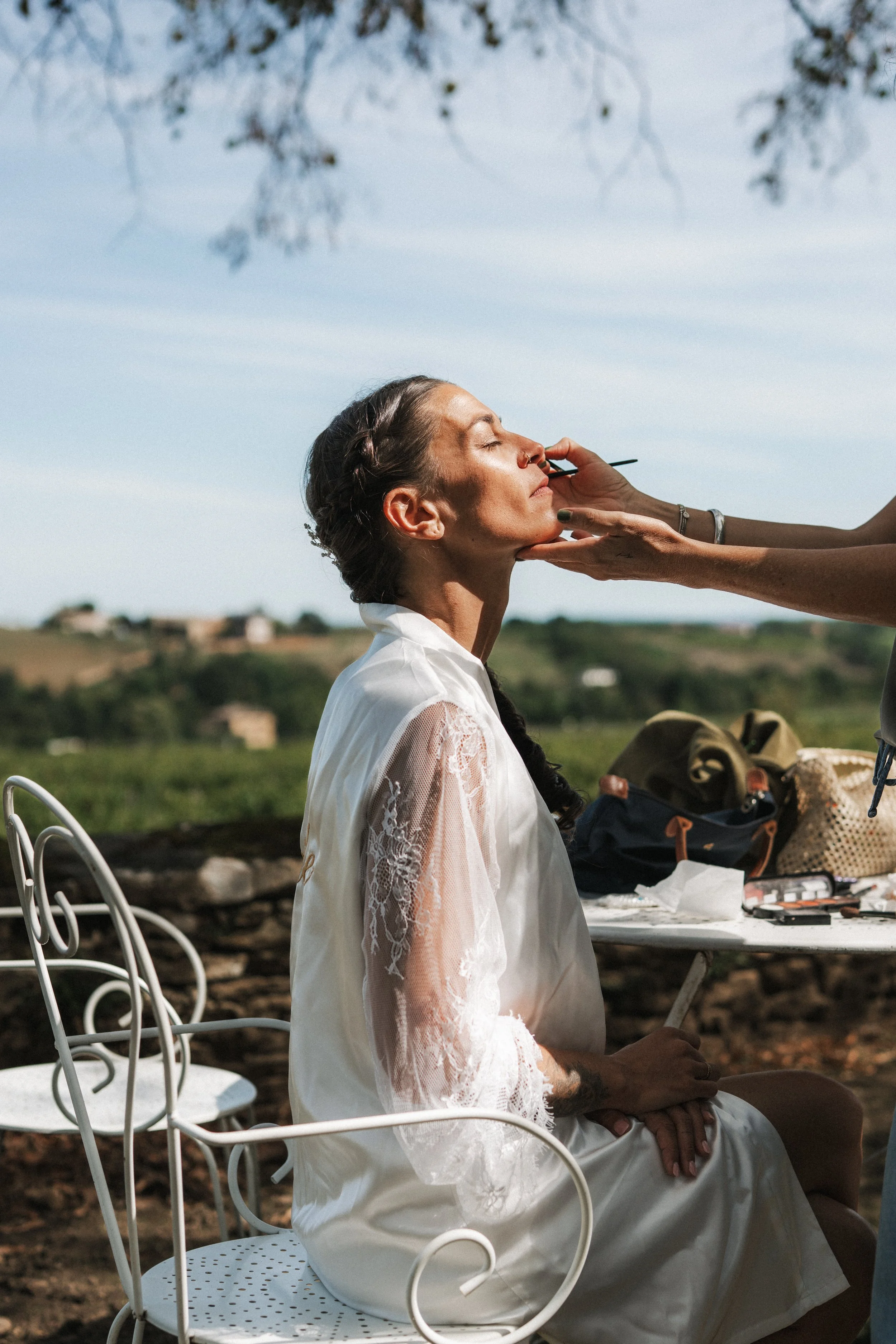 Une femme est en train de se faire maquiller à l'extérieur, avec un ciel bleu en arrière-plan et des collines verdoyantes.