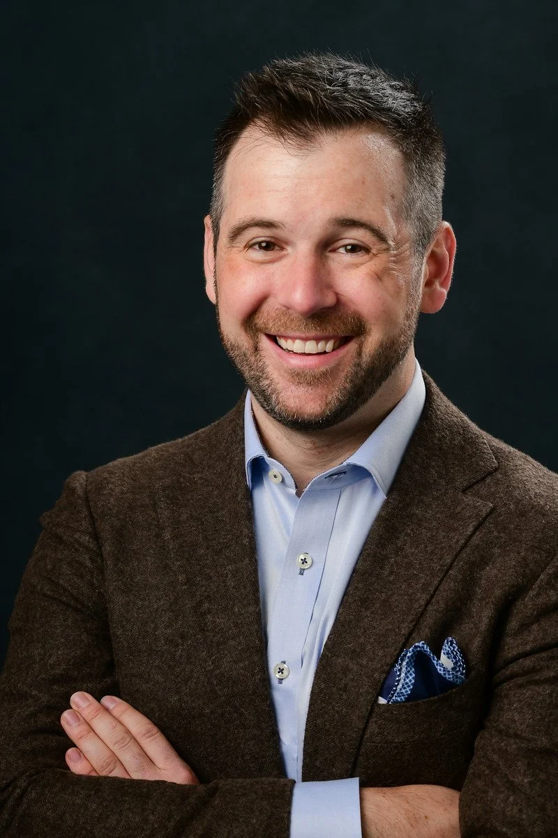 Portrait of a smiling Kevin George Miller with arms crossed, wearing a brown blazer, light blue dress shirt, and a blue patterned pocket square, against a dark background.