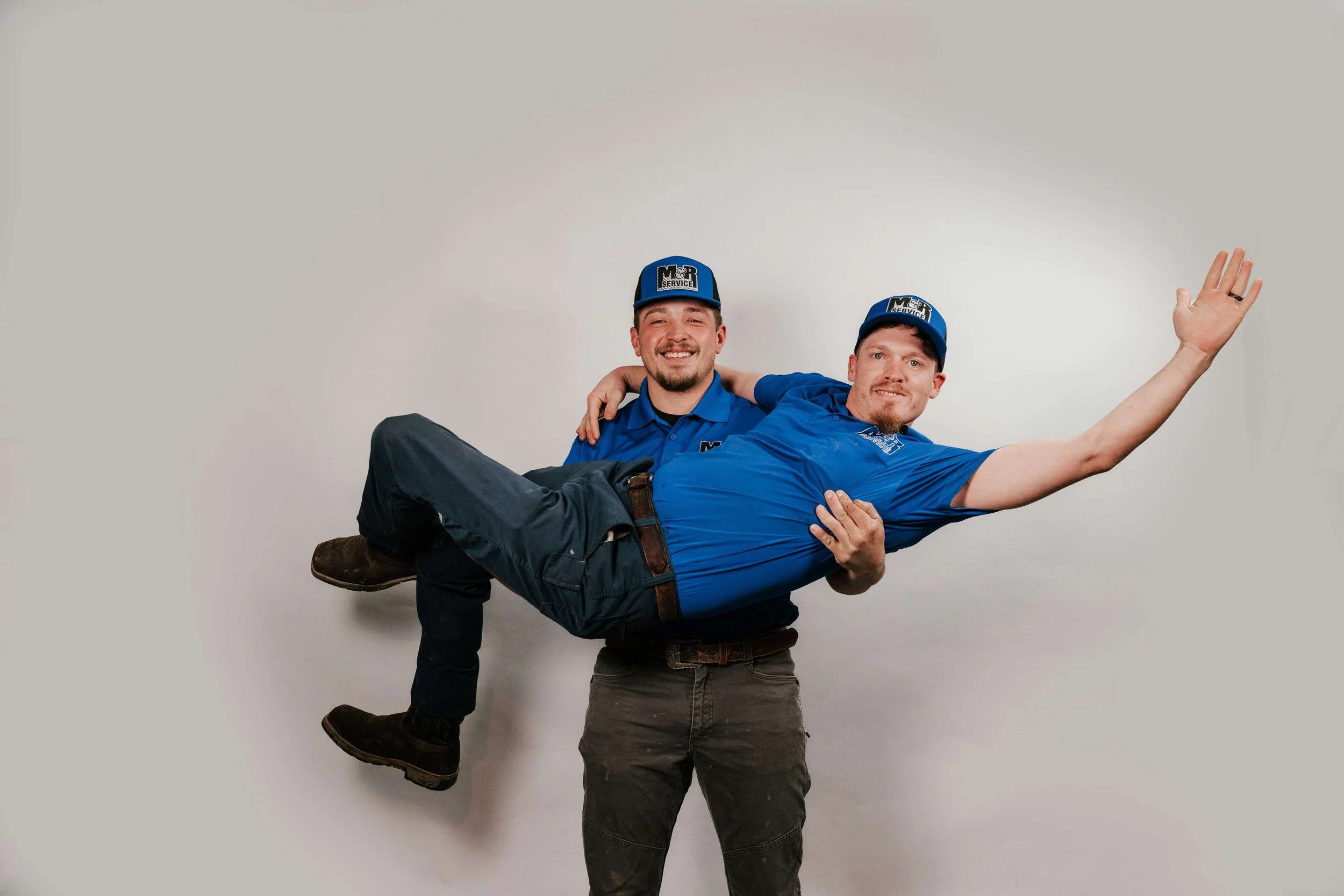Two men in work uniforms, carrying a third man horizontally. All three are smiling and wearing blue caps and shirts with a company logo. The background is plain white.