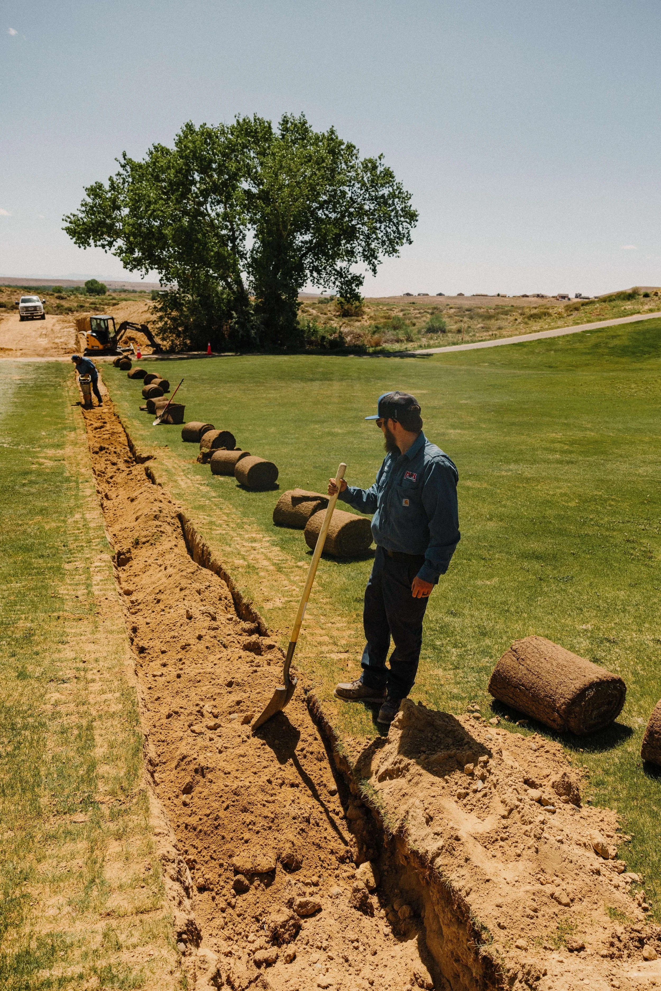 Workers installing underground irrigation pipes in a trench alongside a grassy field with a large tree and construction equipment in the background.
