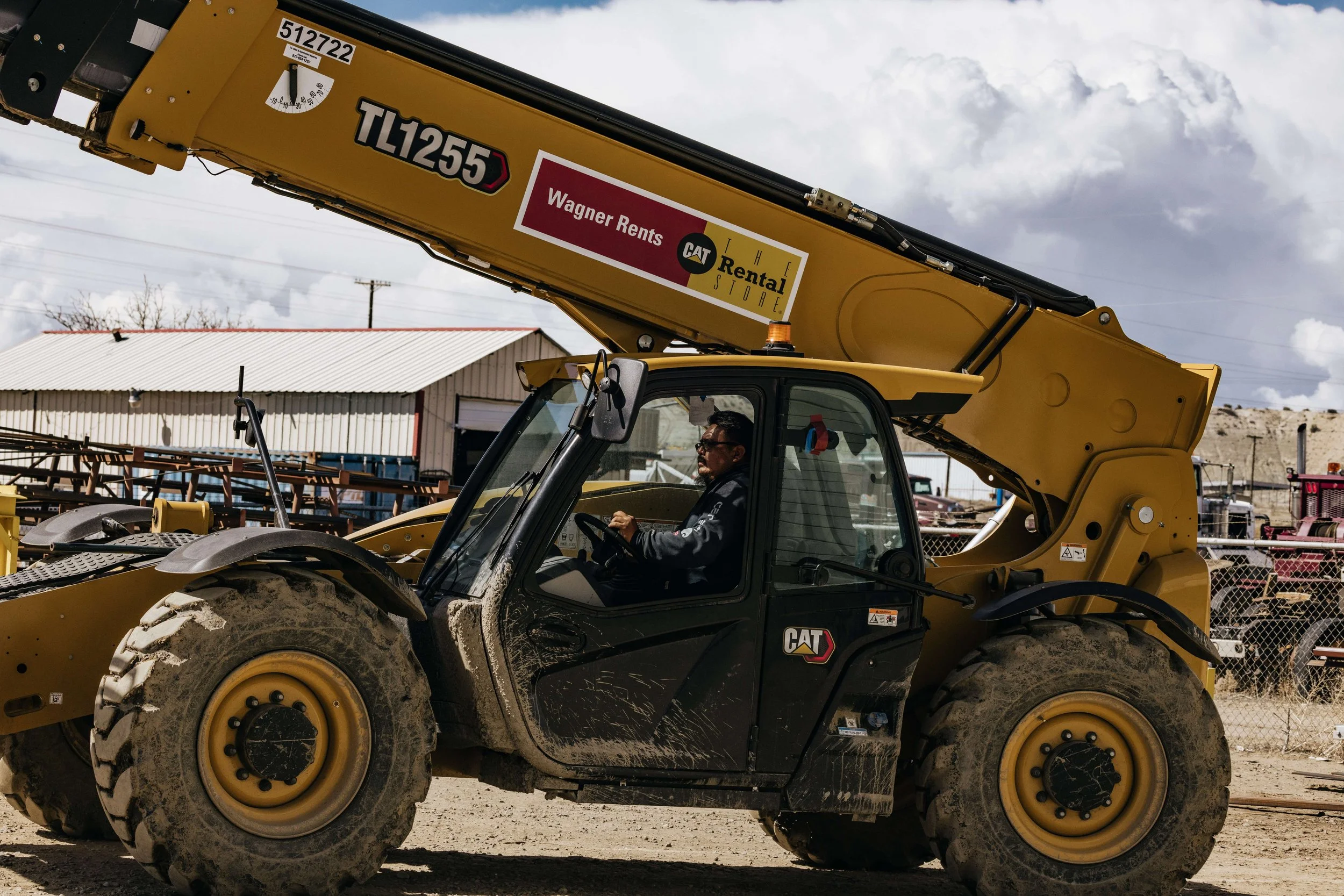 A yellow CAT telehandler with large rugged tires is in a construction or industrial yard. A man is driving the machine, which is parked on a dirt surface, and there are various equipment and a building in the background.