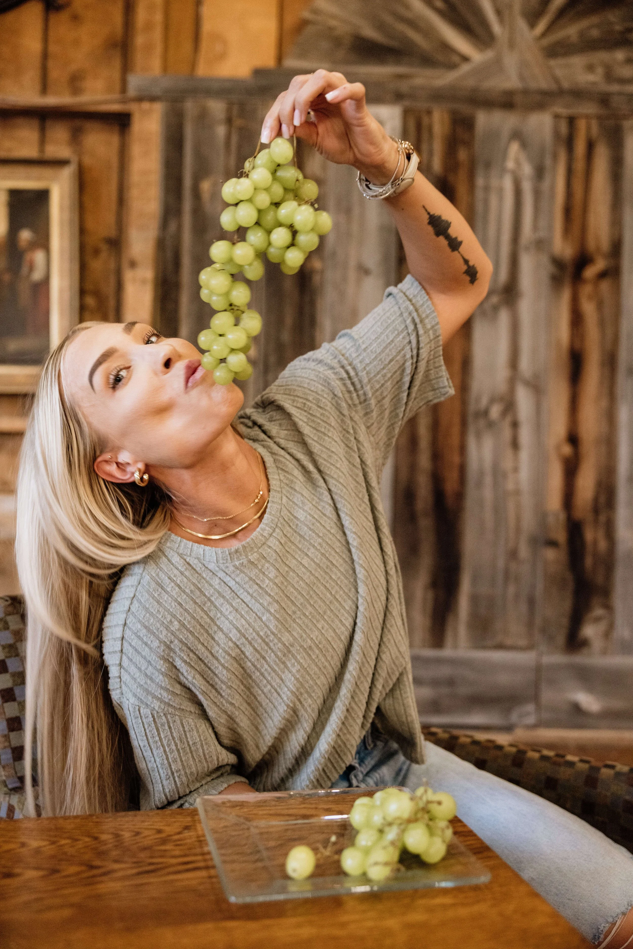 A woman with long blonde hair, light skin, and tattoos is sitting at a wooden table, holding a bunch of green grapes above her mouth as if about to eat them, in a rustic room with wooden walls and frames.