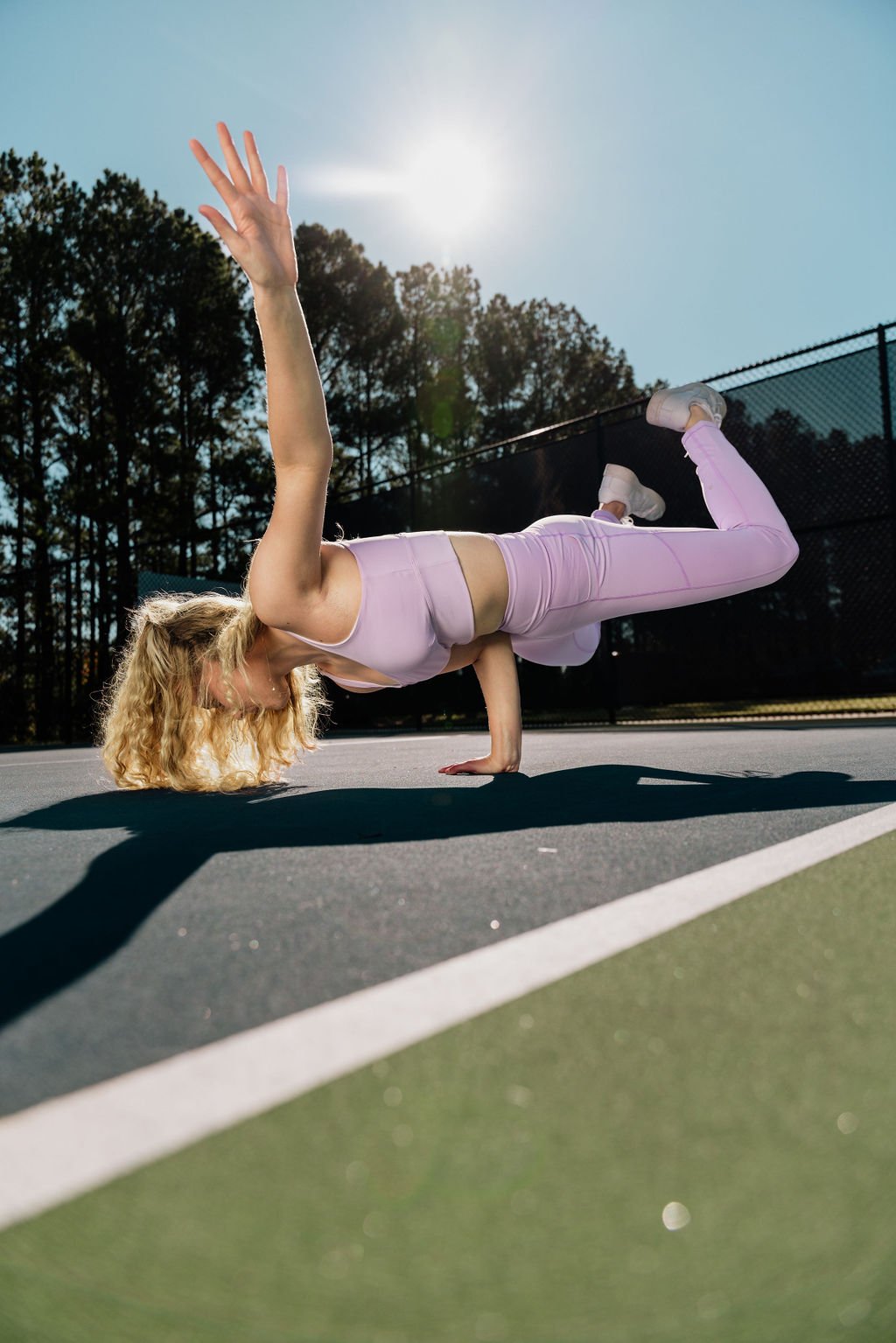 Woman in pastel pink workout clothes performing a one-handed handstand on outdoor tennis court during daytime.