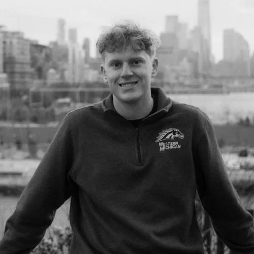 Young man with curly hair smiling, wearing a dark quarter-zip jacket with 'Western Michigan' logo, standing outdoors with an urban cityscape in the background.