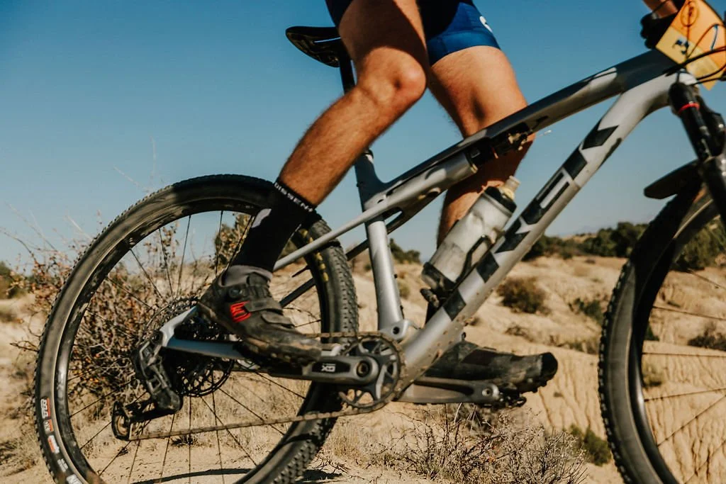 Close-up of a person riding a Trek mountain bike through a desert landscape with dry bushes and sandy terrain.