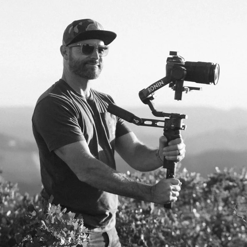 Man wearing sunglasses, cap, and casual t-shirt, holding a camera stabilizer with a camera attached, standing outdoors with a blurred landscape in the background.