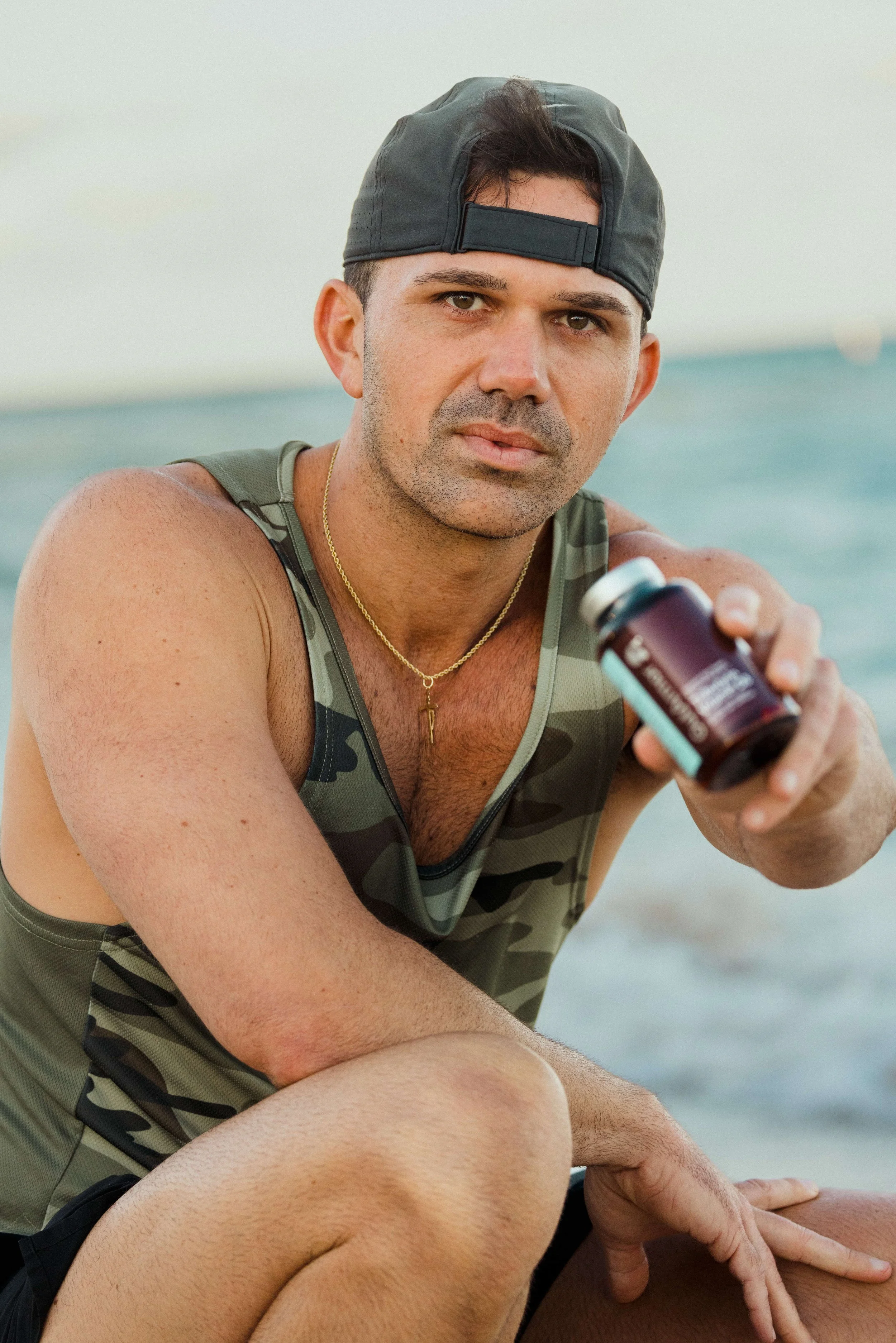 A man with a serious expression on a beach, holding a supplement bottle and wearing a camouflage tank top and a backward cap.
