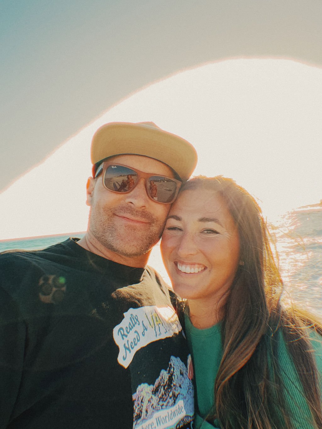 A man and woman smiling on a beach at sunset, with the man wearing sunglasses and a cap and the woman with long brown hair.