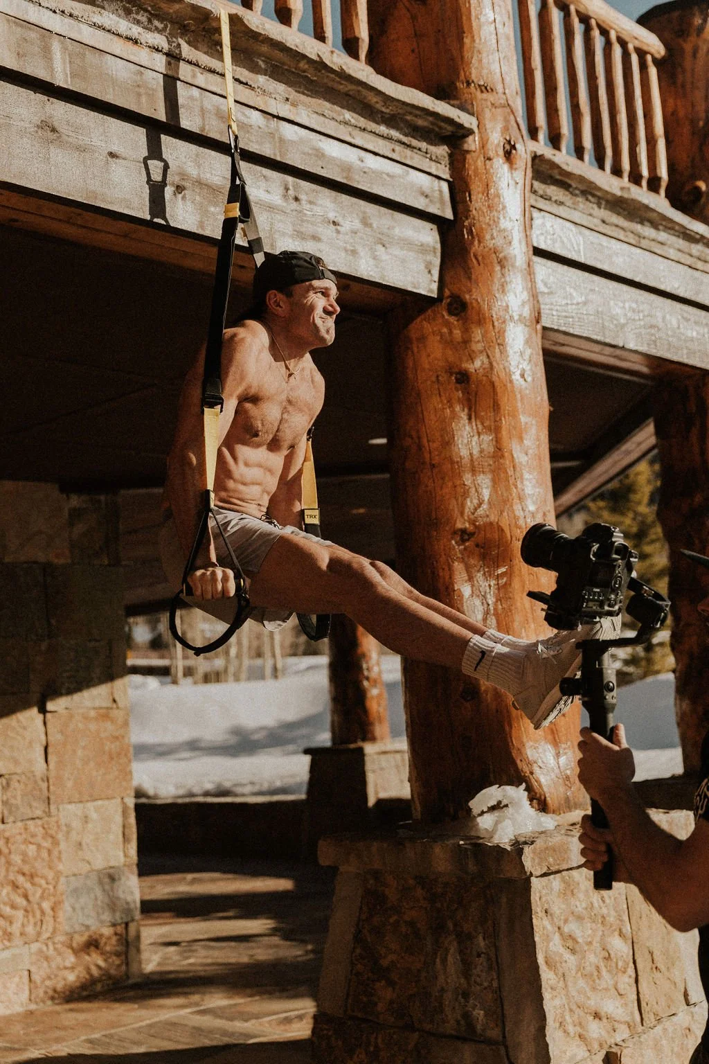 A shirtless man doing an outdoor workout hanging from gym straps, with a camera recording him, in a rustic wood and stone setting.