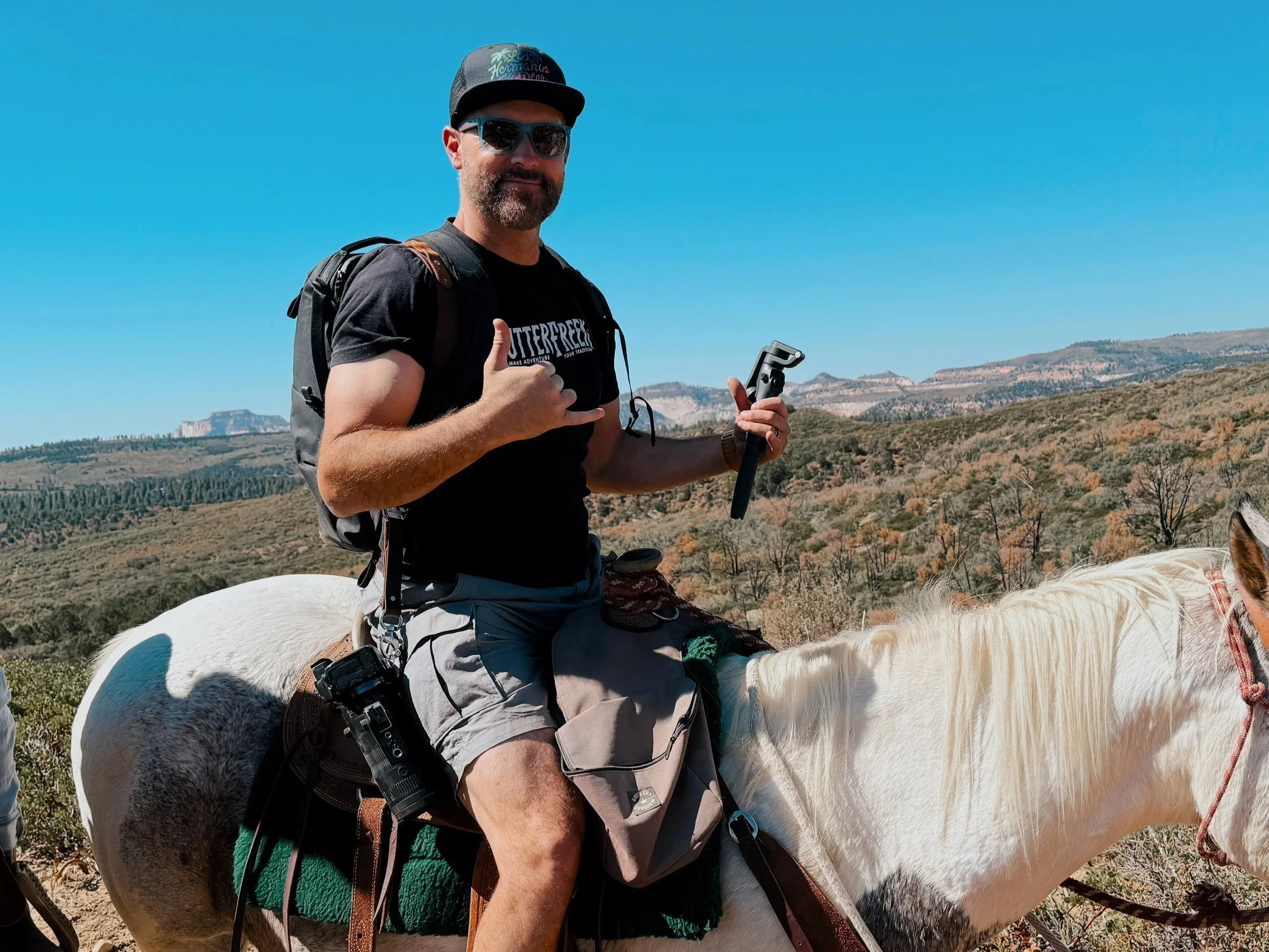 A man with sunglasses, a black T-shirt, and a baseball cap riding a white horse in a desert landscape with mountains in the background. He is making a shaka sign with his right hand and holding a camera stabilizer in his left hand.