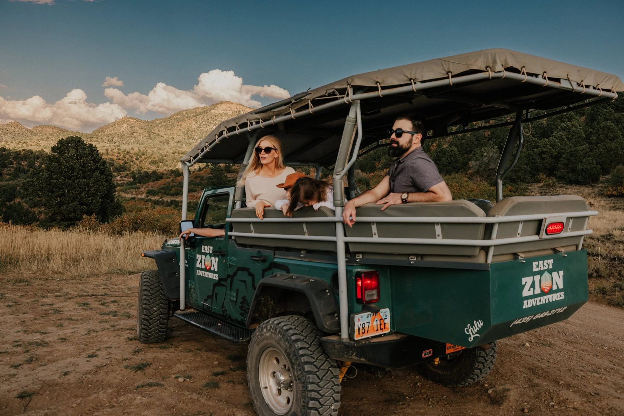 Three people and a dog on an open-air safari vehicle in a mountainous landscape during daytime.