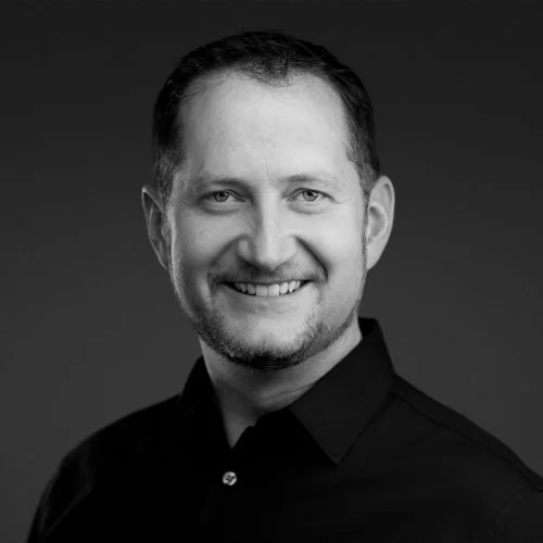 Black and white portrait of a smiling man with short dark hair and a beard, wearing a dark button-up shirt.