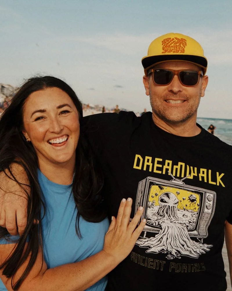 A smiling woman with long dark hair wearing a blue top, and a man wearing sunglasses, a yellow cap, and a black T-shirt with a graphic and text, at the beach.