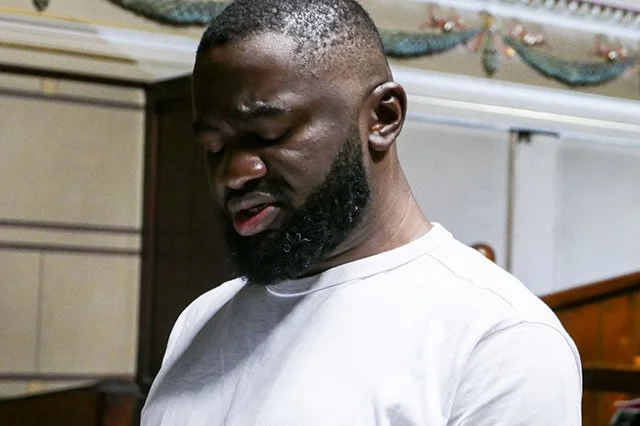 A man with a beard wearing a white t-shirt looking down in an indoor setting.