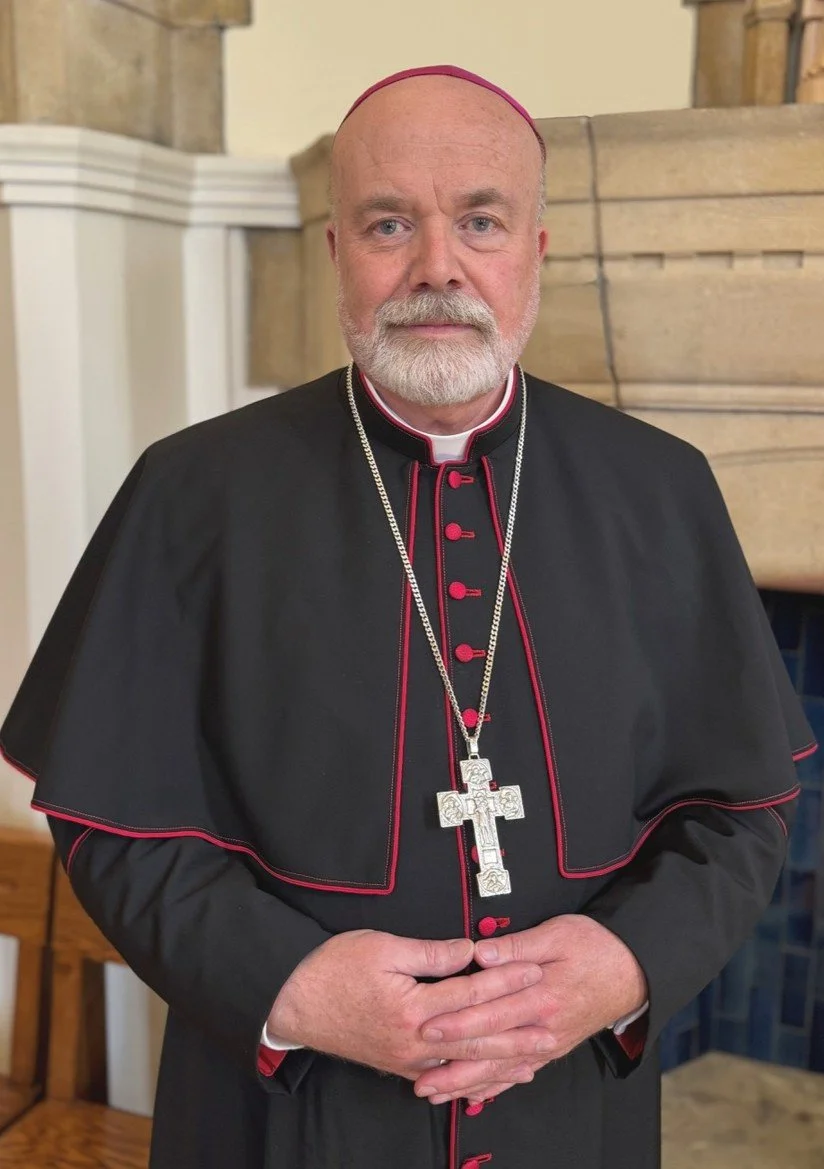 Bishop Marcus Stock wearing a black cassock with red piping, a silver chain with a large cross pendant, and a pink skullcap, standing indoors in front of a stone and wooden background.