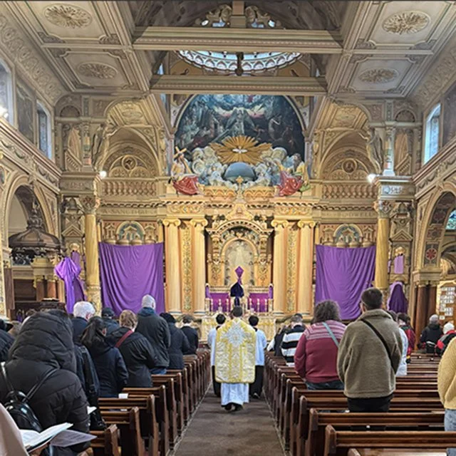 People attending a church service or ceremony in a grand, ornate church with intricate ceiling designs, large purple drapes, and a mural above an altar. The clergy are dressed in gold and white robes, facing a person on a purple throne-like chair.