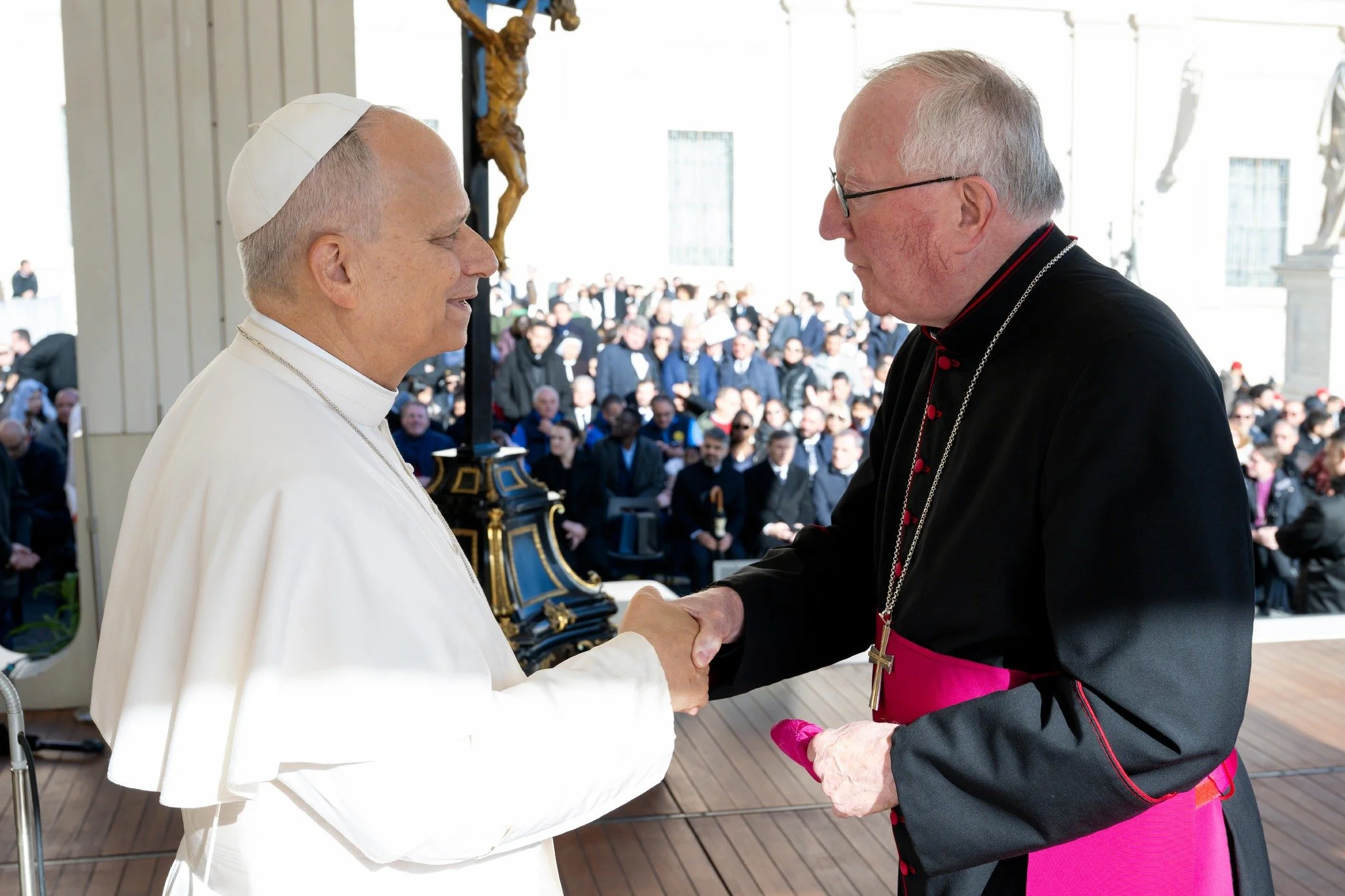 Pope Francis and a Catholic bishop shaking hands during an outdoor religious ceremony, with a crowd of people in the background.