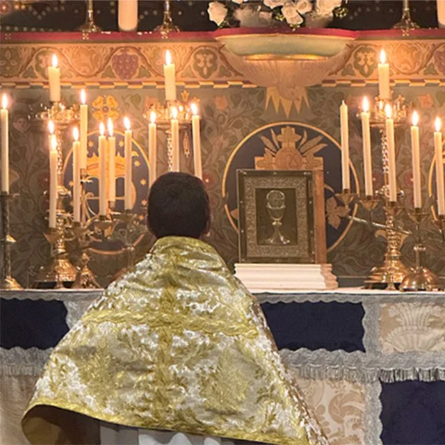A person with short dark hair, draped in a gold and white ceremonial cloth, kneels before a religious altar adorned with lit candles, ornate candles holders, and religious icons.