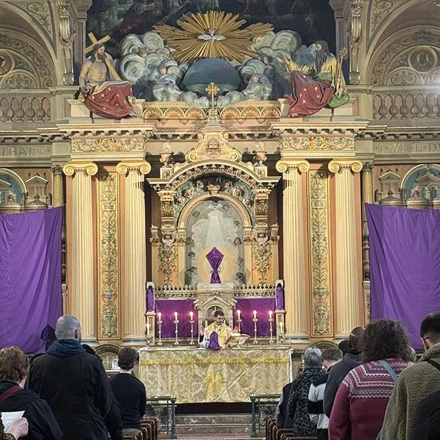 Interior of a church with an ornate altar decorated with purple cloth and lit candles, congregation gathered facing the altar, with large purple curtains on either side, and a religious painting with figures and clouds above.