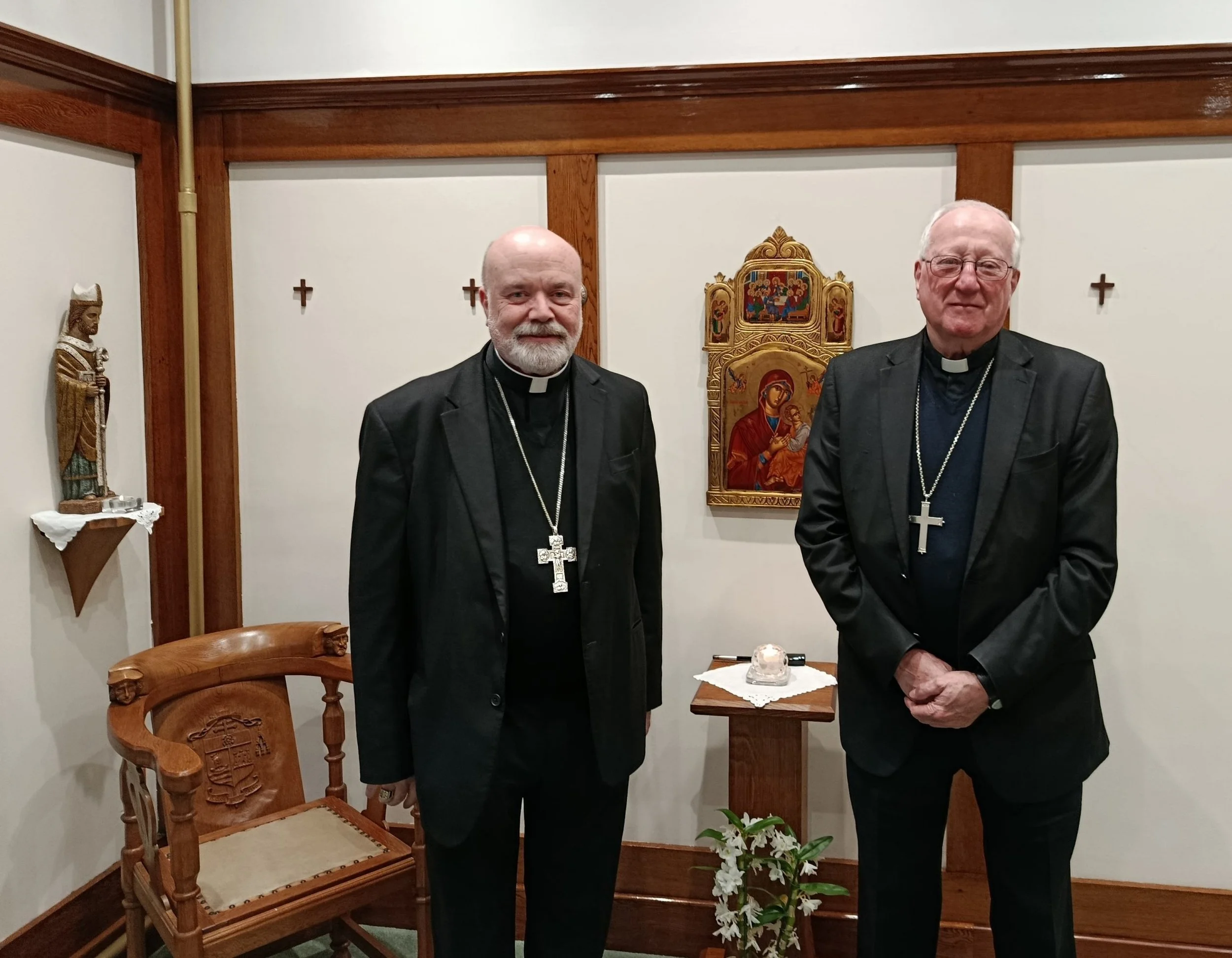 Two clergymen standing in a room with religious icons on the wall, wearing black suits and cross necklaces, with a wooden chair and religious statues decor.