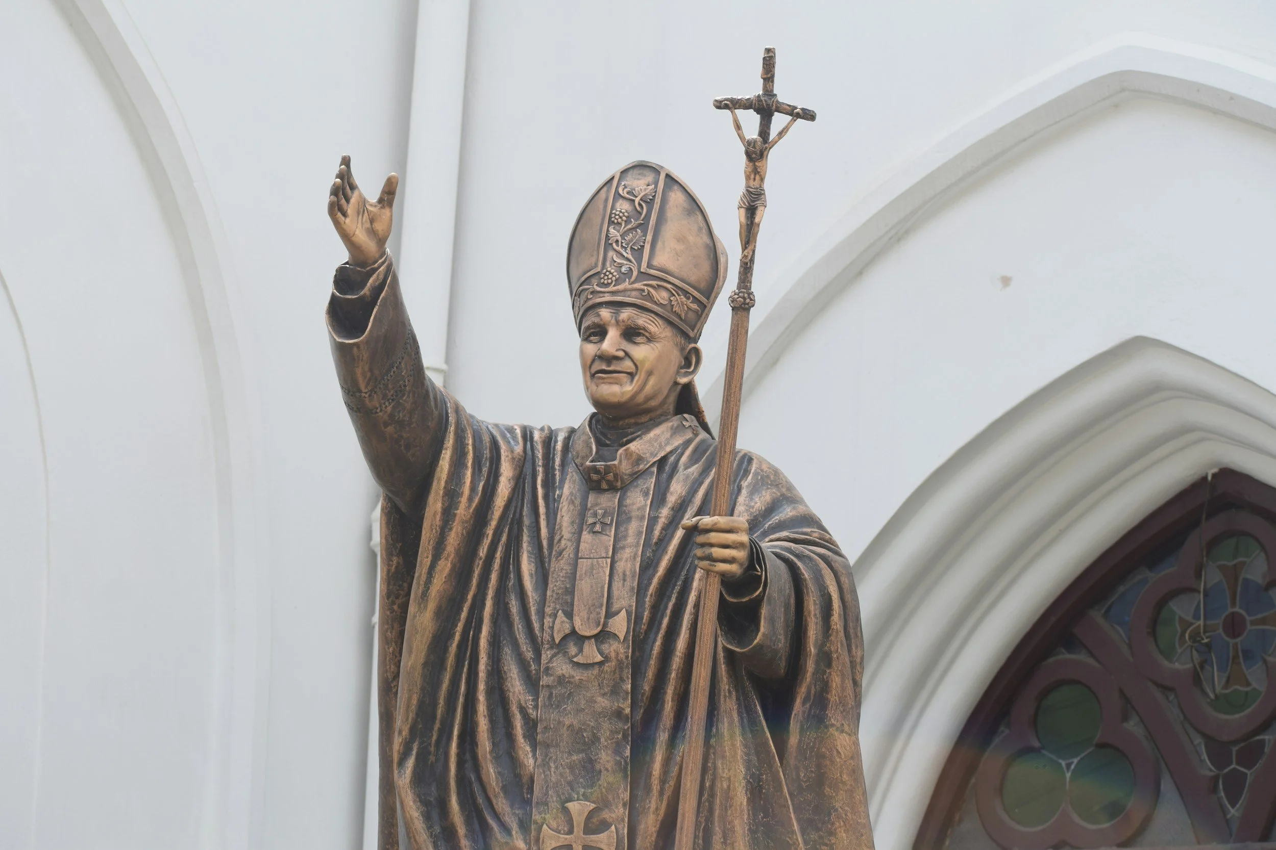 A bronze statue of a bishop or pope figures outside a church, holding a staff with a crucifix and raising one hand in blessing.