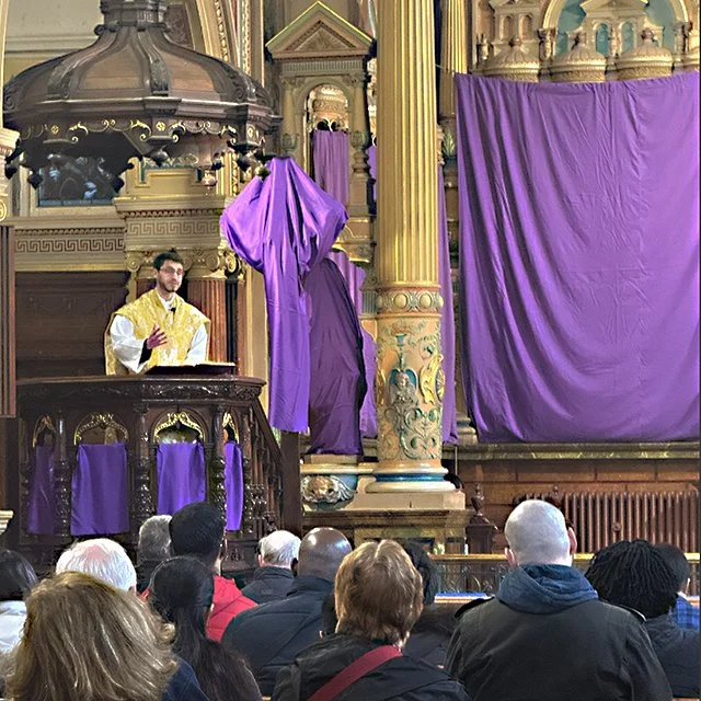 A man dressed in a yellow and white robe standing at a pulpit in a church with purple drapes covering the altar and decorations, and a congregation seated facing the pulpit.