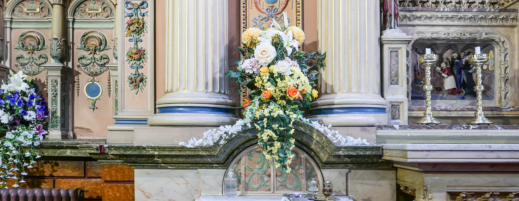 A floral arrangement with white, yellow, and orange flowers on a decorated altar at Saint Charles Borromeo church in Hull UK, with detailed columns, religious artwork, and candlesticks in the background.