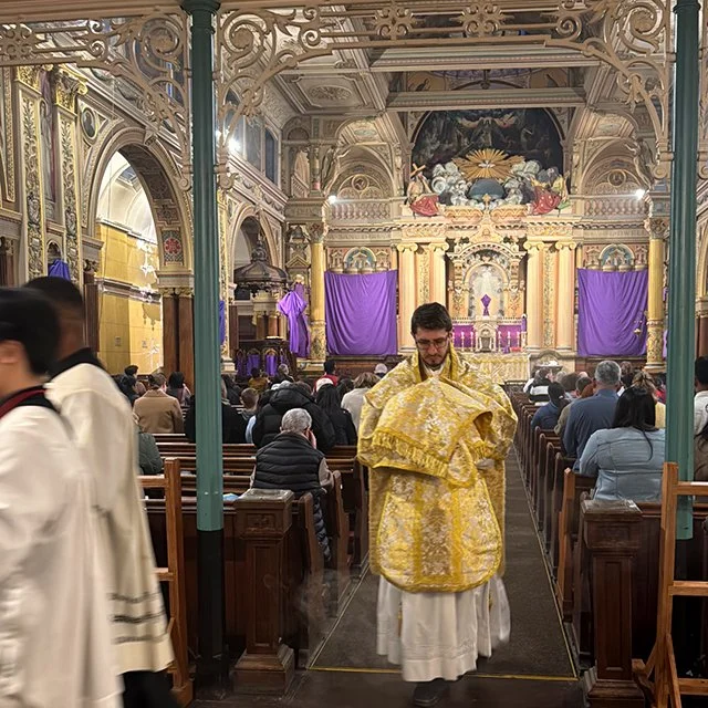 A priest in ornate gold vestments walks down the center aisle of a church during a service, with worshippers seated in pews on both sides and an elaborate altar in the background.