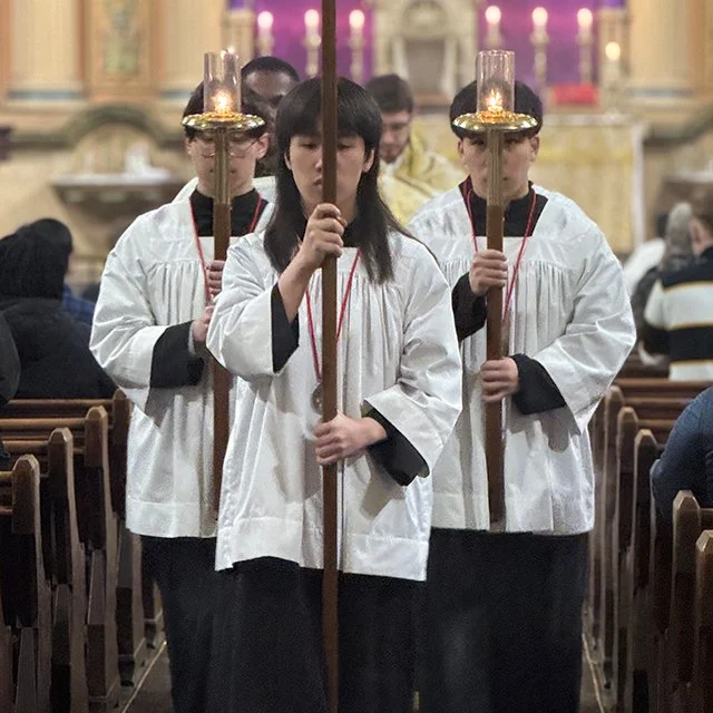 Four children in white robes standing in a church, holding candles with glass covers and a tall candle holder, participating in a religious ceremony.