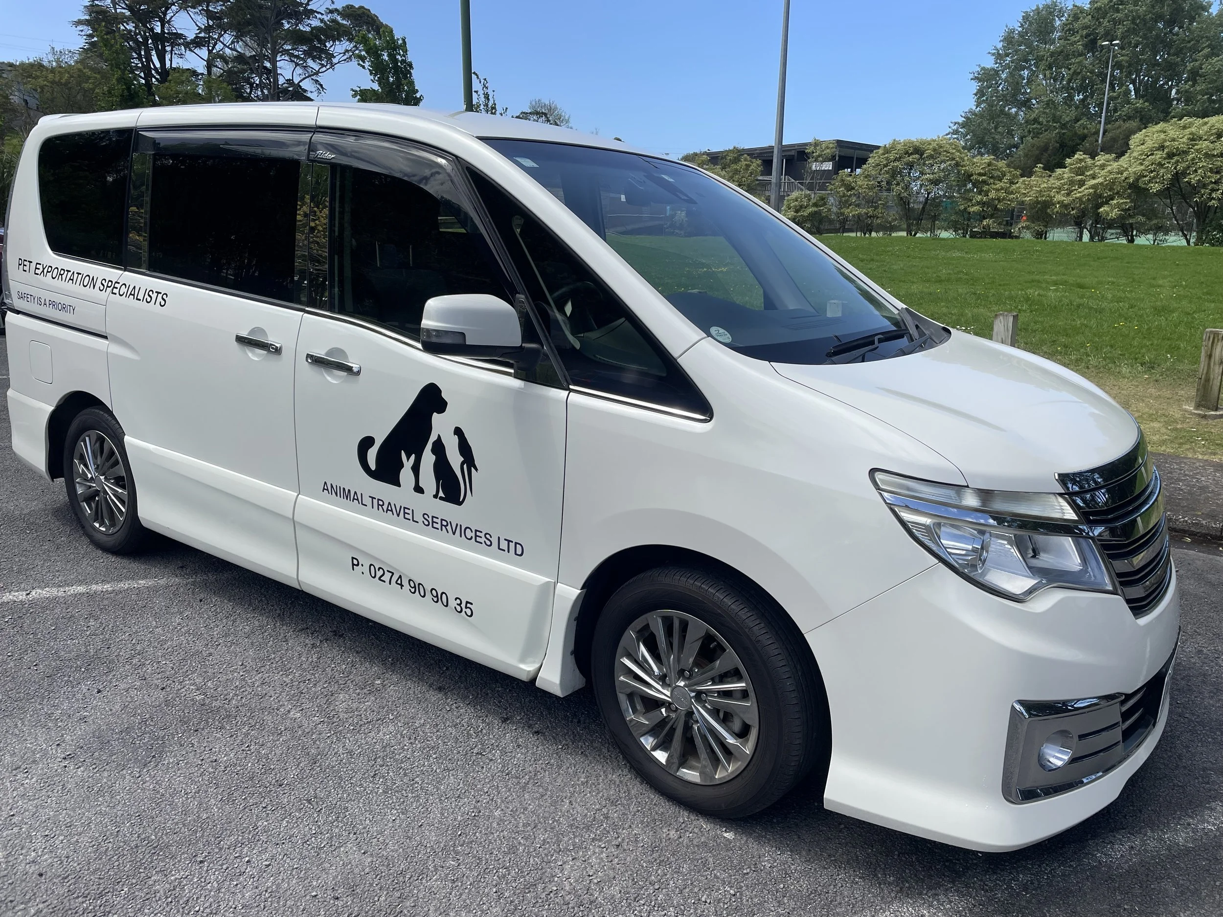White van with black animal silhouettes and text for pet travel services parked on the street near a grassy park with trees.