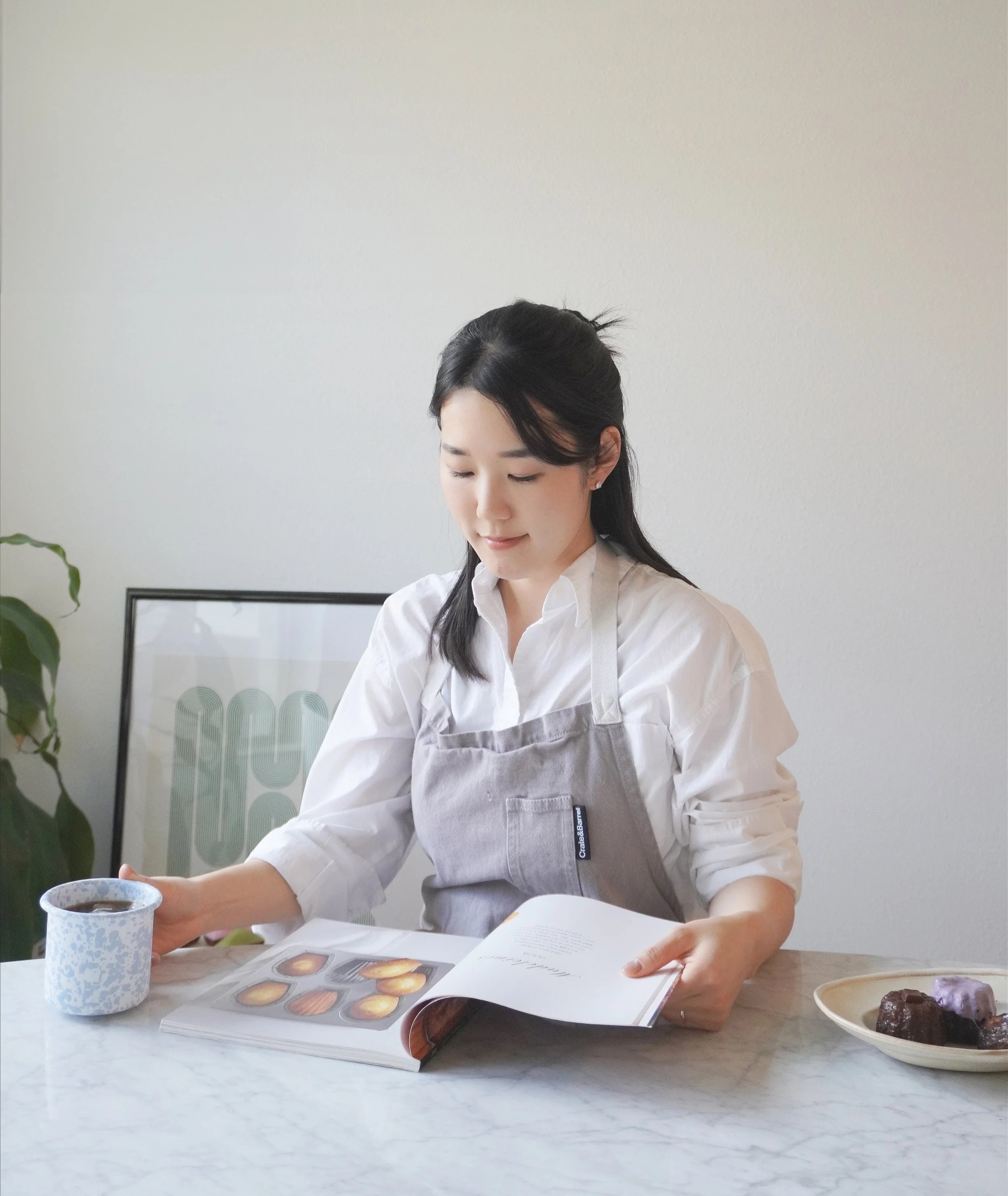 A woman sitting at a table with a book, a cup of coffee, and a plate of assorted pastries, wearing a white shirt and gray apron, looking at the book.