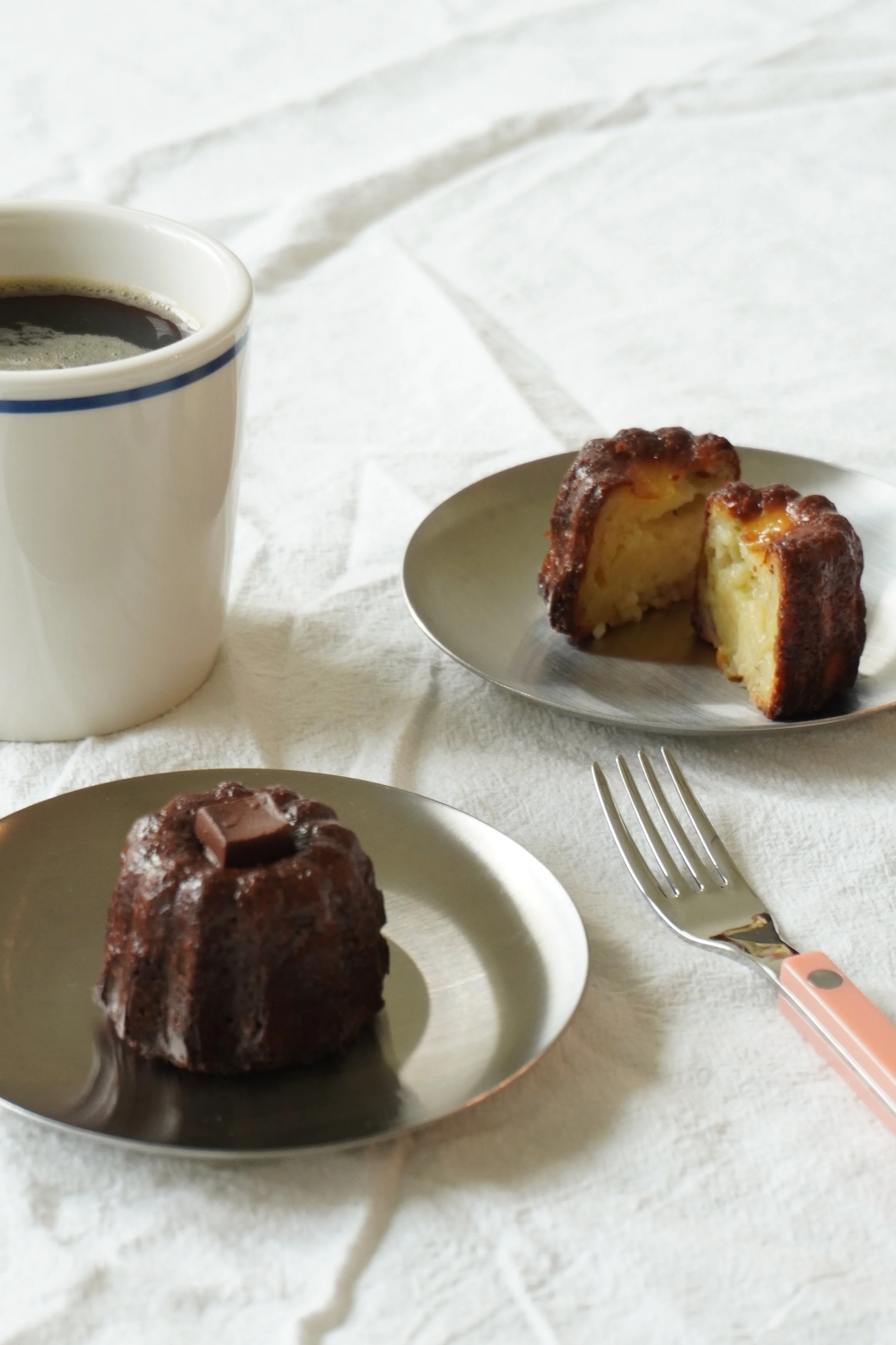 Chocolate bundt cake with a chocolate piece on top, chocolate coffee in a white mug, and a slice of a different cake on a small plate on a white cloth surface.
