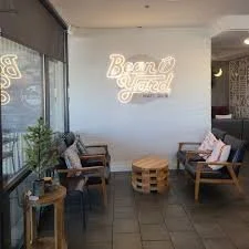 Indoor seating area with wooden benches and a table made from wooden pallets, a neon sign on the wall reads 'Bean & Brew,' and there are some plants and cushions for comfort.
